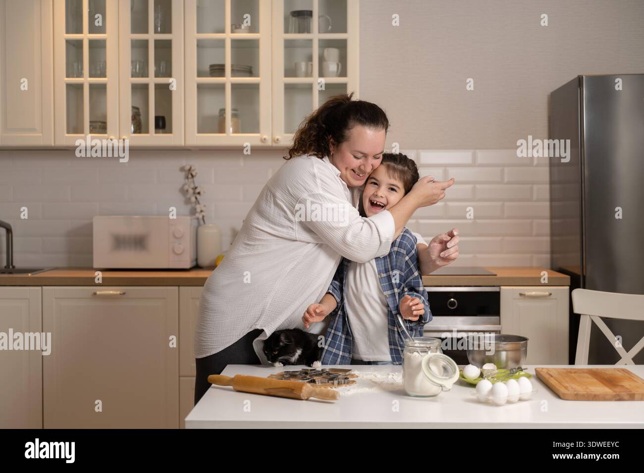 Eine fröhliche Mutter umarmt ihre Tochter, während sie gerne zusammen backen und unvergessliche Momente in ihrer gemütlichen Küche schaffen. Mehl und Zutaten sind Stockfoto