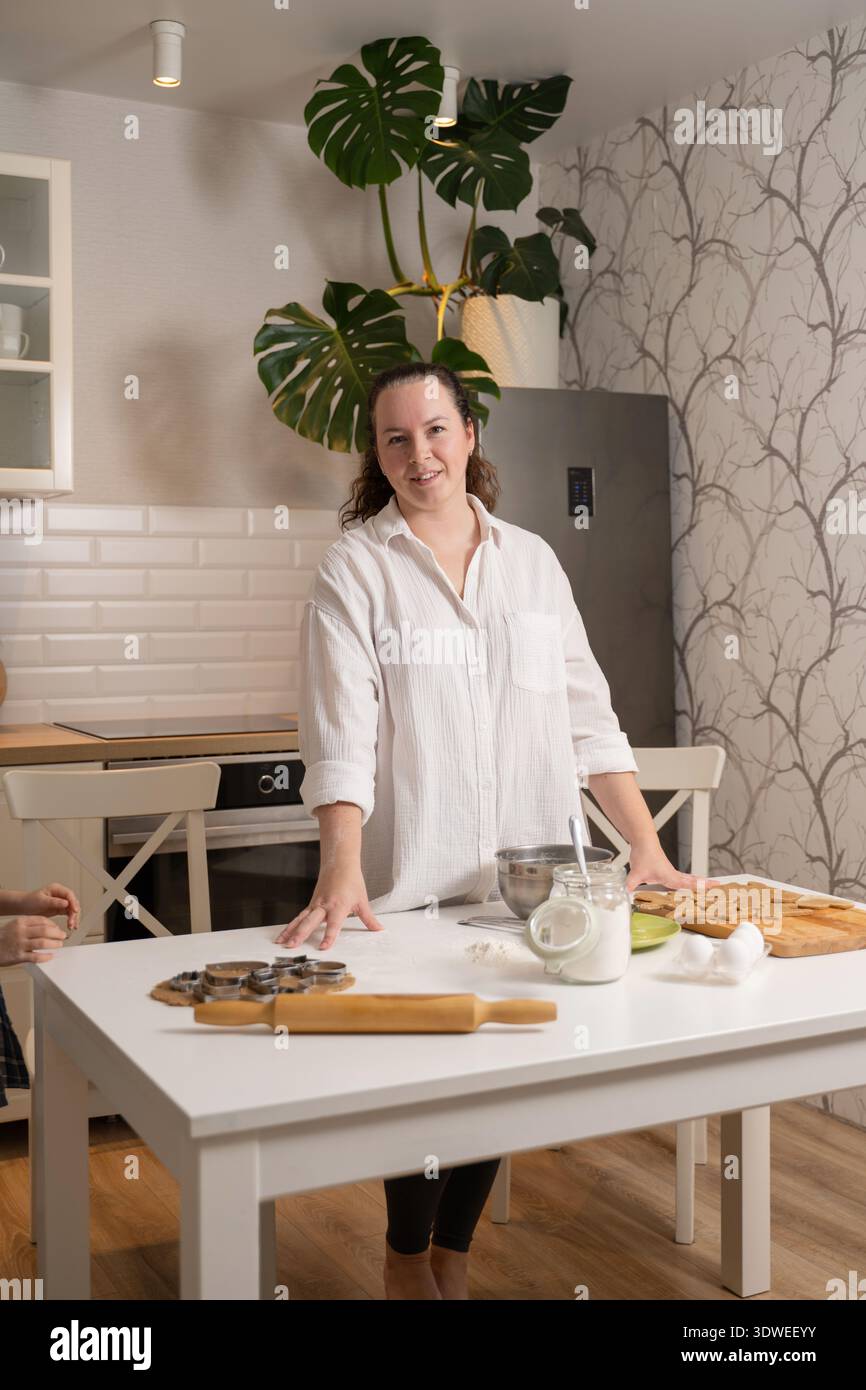 Eine Frau in einer modernen Küche bereitet hausgemachte Kekse zu. Zutaten und Backwerkzeuge sind ordentlich auf dem Tisch angeordnet, was eine einladende und gemütliche Atmosphäre schafft Stockfoto