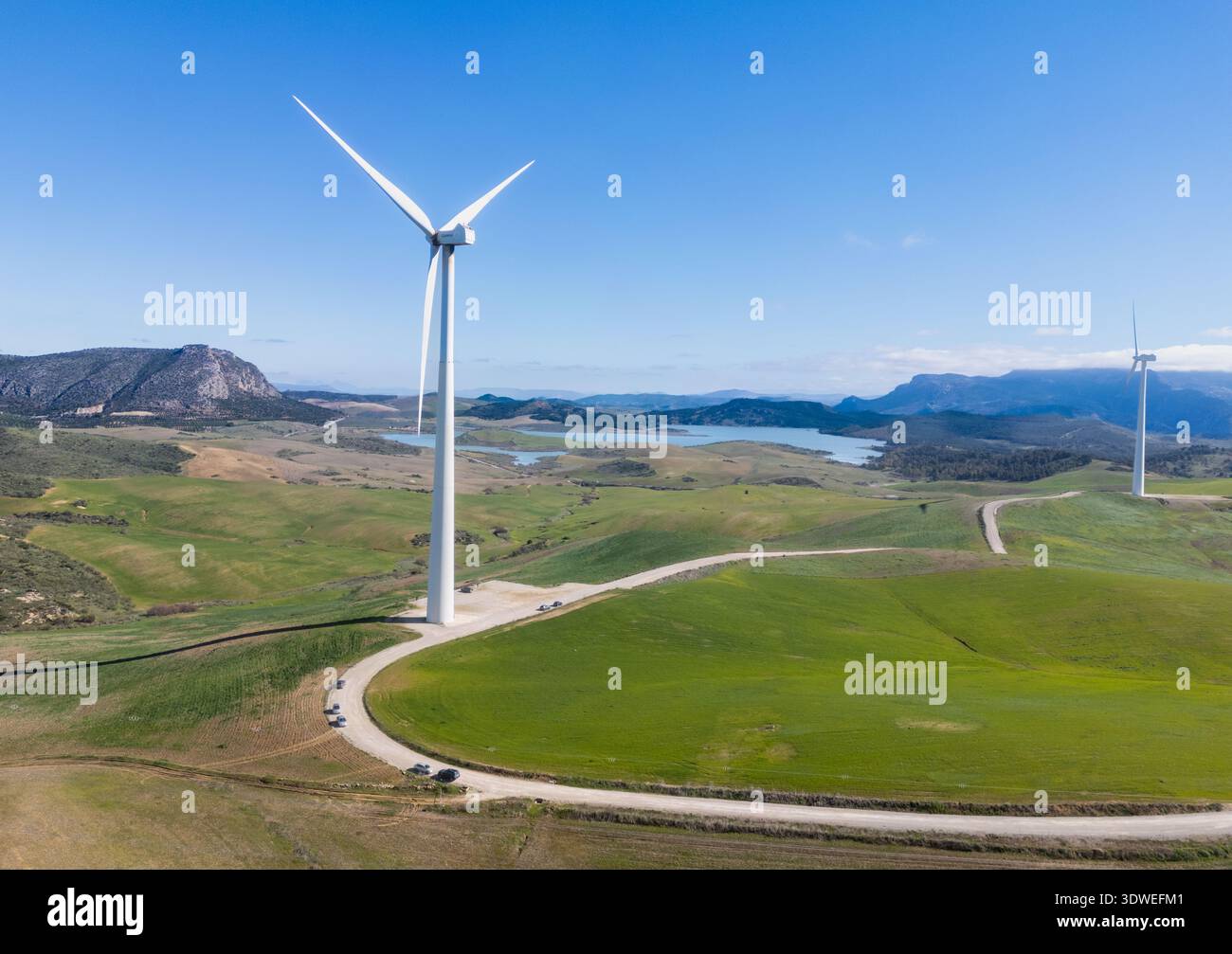 Aus der Vogelperspektive von Windturbinen, die hoch stehen vor einem Hintergrund von sanften grünen Hügeln und blauem Wasser, wodurch ein auffälliger Kontrast von Farbe und Form entsteht; Stockfoto