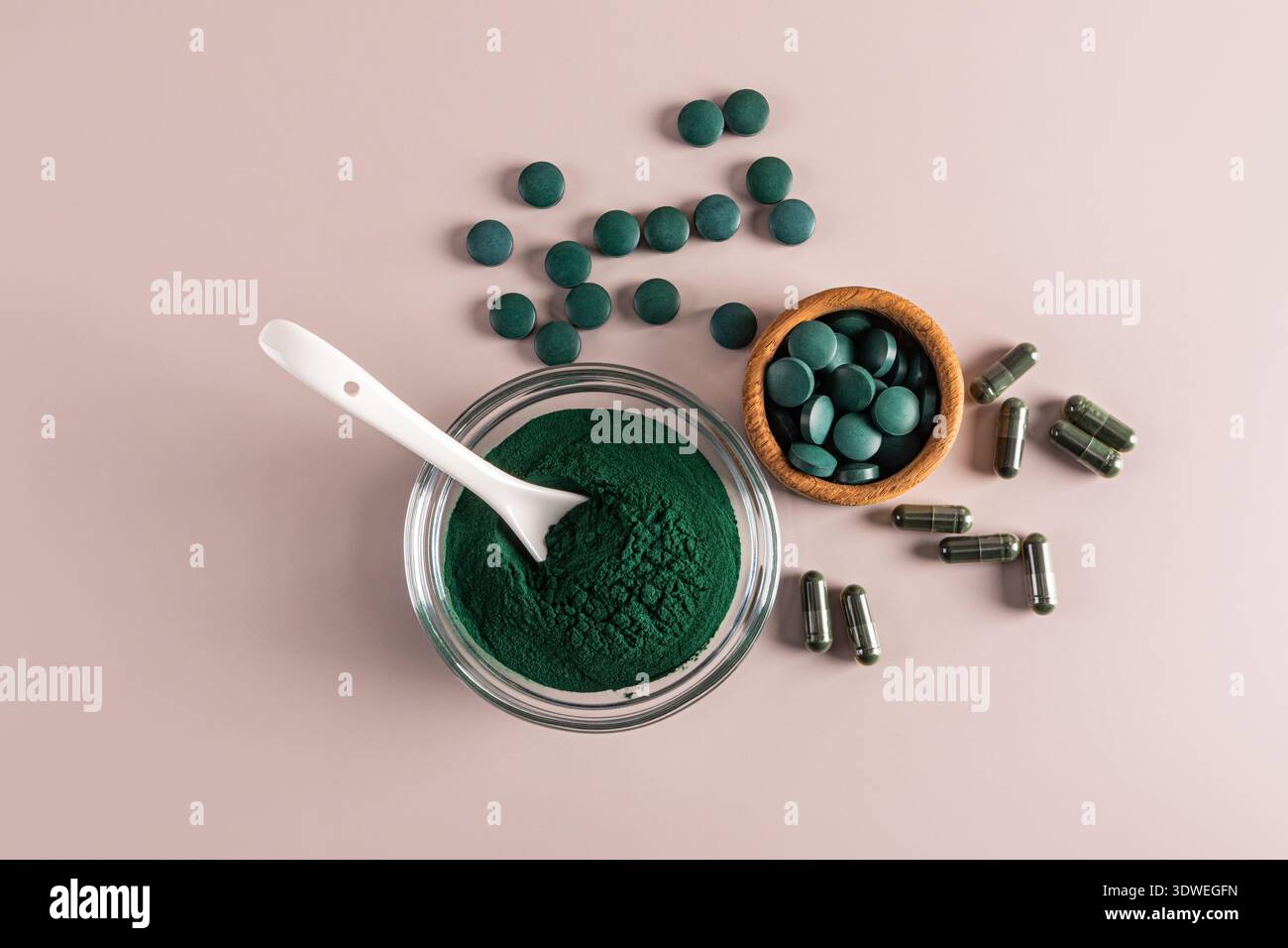 Blick von oben auf Spirulina Pulver in einer Glasschale mit Löffel, umgeben von Spirulina Tabletten und Kapseln. Der Hintergrund ist eine weiche, pastellrosa Oberfläche. Stockfoto