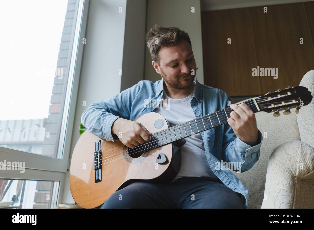 Ein junger Mann spielt lässig eine Akustikgitarre in seiner gemütlichen Umgebung. Er ist konzentriert und entspannt und genießt die beruhigende Atmosphäre am Fenster o Stockfoto