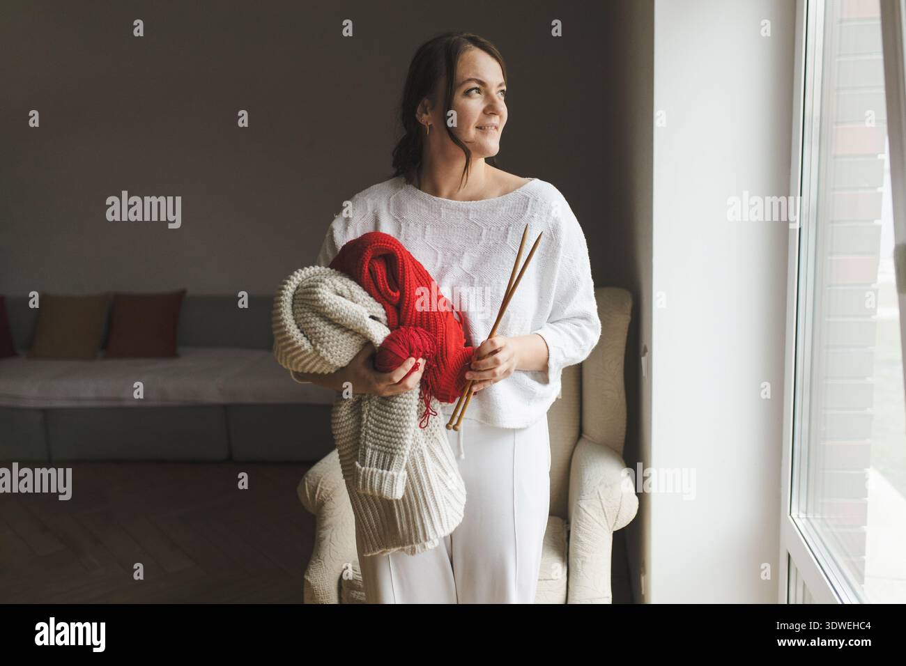 Eine Frau genießt ihr Strickhobjekt, hält Stricknadeln und einen leuchtend roten Pullover. Die gemütliche Szene unterstreicht Kreativität und Entspannung bei Sonnenlicht Stockfoto