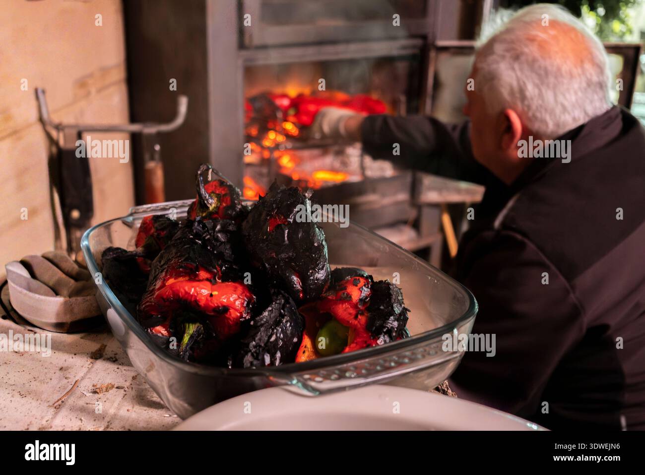 Rückansicht eines nicht erkennbaren älteren Mannes, der rote Paprika über offener Flamme in der rustikalen Küche röstet, wobei traditionelle Kochmethoden und heimeliger geldautomat hervorgehoben werden Stockfoto