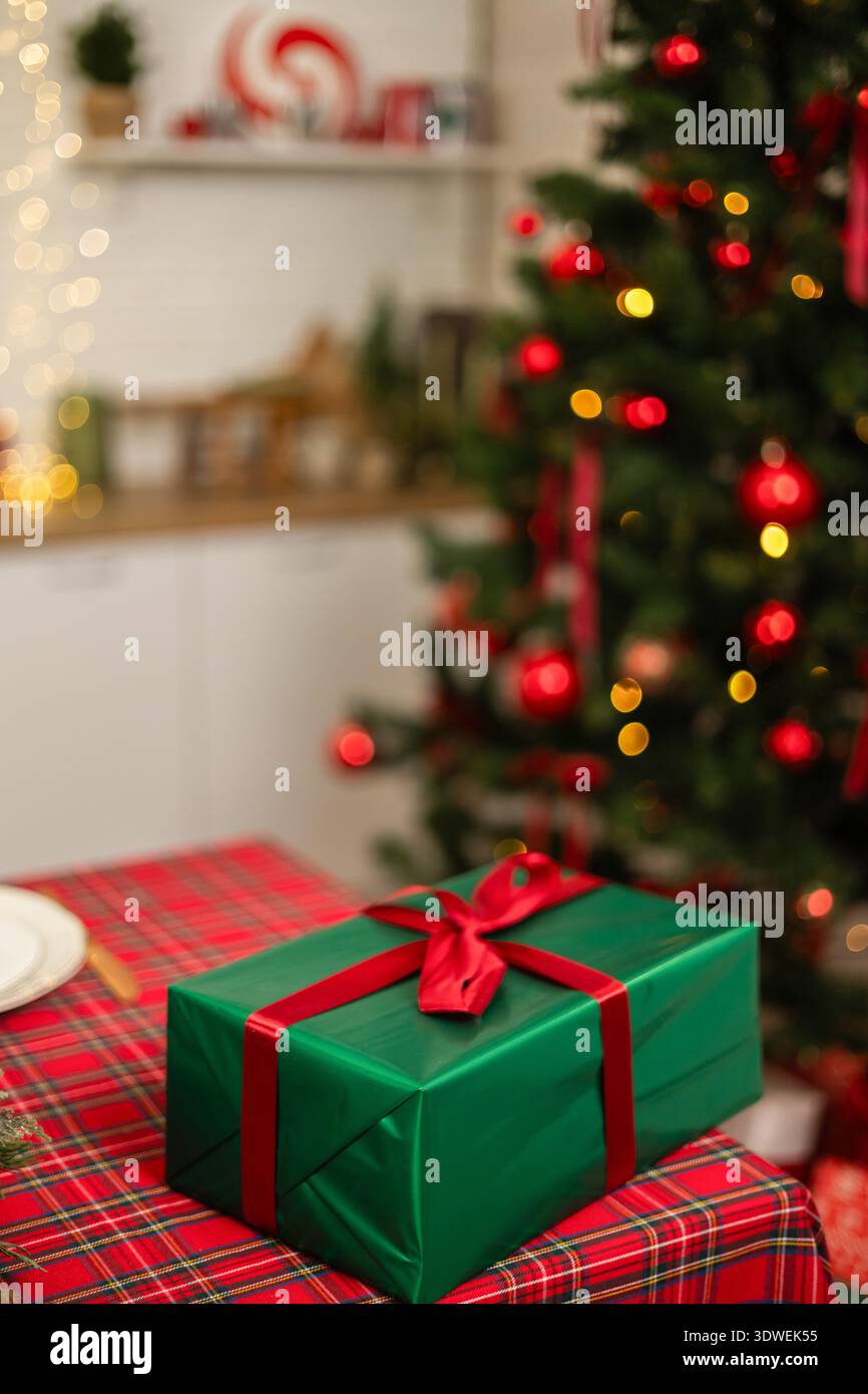 Ein wunderschön gewickeltes, grünes Geschenk mit rotem Band sitzt auf einer karierten Tischdecke vor einem leuchtenden Weihnachtsbaum, der mit roten Ornamenten und warmen Farben verziert ist Stockfoto
