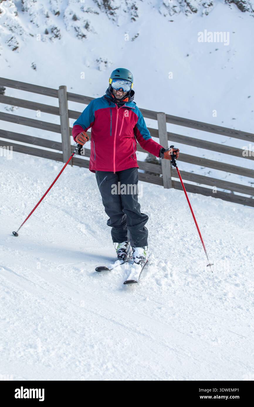 Ein Mann in Skiausrüstung steht selbstbewusst auf einer verschneiten Piste und hält Skistöcke. Er trägt eine farbenfrohe Winterjacke, Helm und Schutzbrille, umgeben von einem Winter Stockfoto