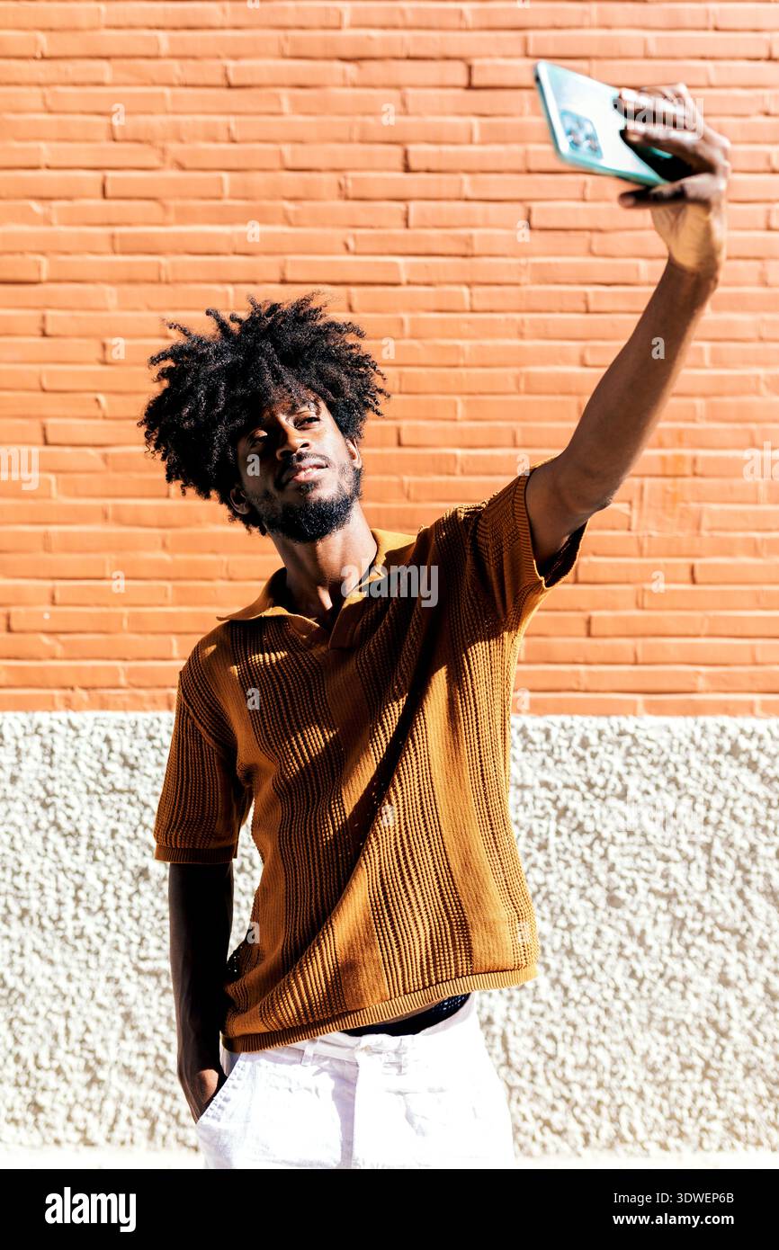Ein Mann mit Afro-Frisur macht ein Selfie gegen eine sonnendurchflutete orangene Ziegelwand. Er trägt ein braunes strukturiertes Hemd und wirkt entspannt und selbstbewusst Stockfoto