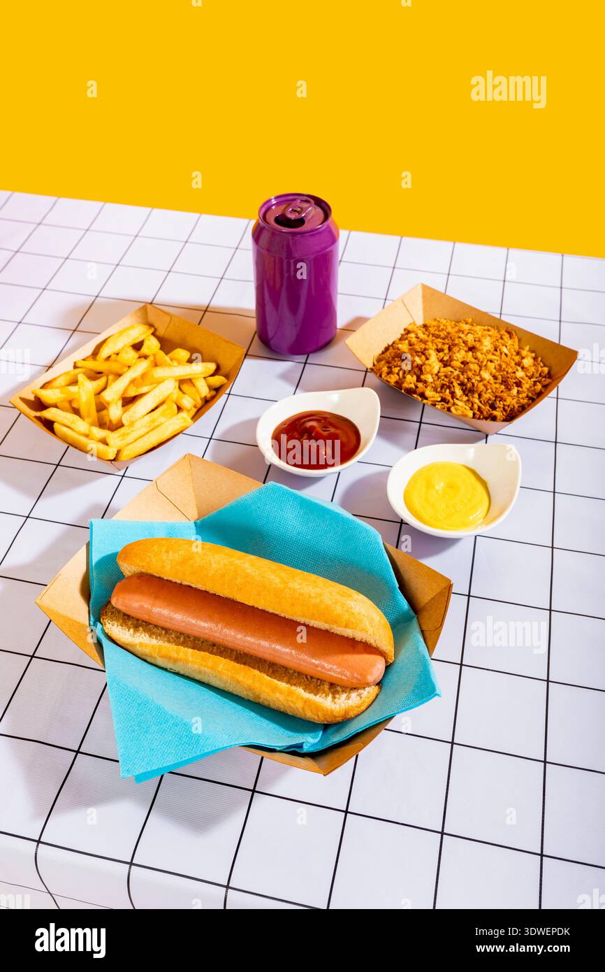 Eine lebhafte Mahlzeit mit einem Hot Dog in einem Brötchen auf einer blauen Serviette, dazu knusprige Pommes Frites, eine Schüssel knuspriger Snacks und eine lila Dose, alles auf einem Stockfoto