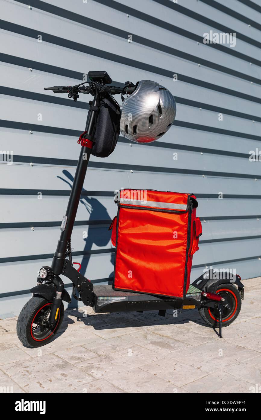 Elektroroller, der von einem Fahrer verwendet wird, mit einer hellroten Liefertasche und einem Helm, der in einer städtischen Umgebung an einer modernen Gebäudewand geparkt wird Stockfoto