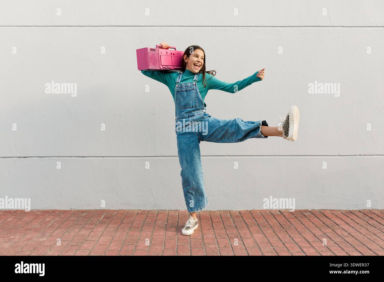 Ein fröhliches Mädchen in Denim-Overalls und einem grünen Pullover tanzt mit Begeisterung und hält eine pinkfarbene Boombox. Ihr fröhlicher Ausdruck fängt das Wesen ein Stockfoto