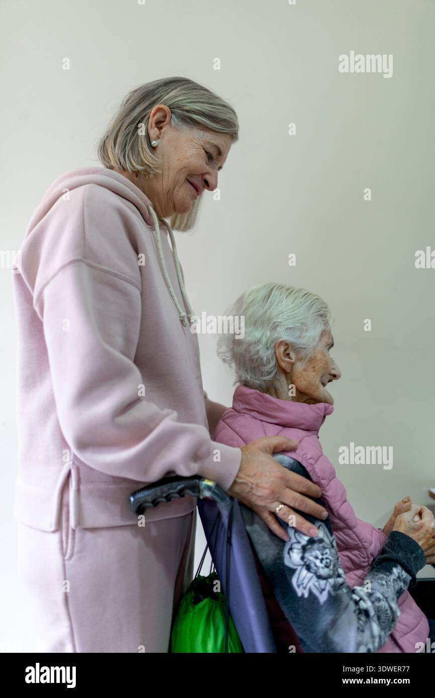 Eine ältere Frau in einem Seniorenwohnheim wird in Pflege gezeigt. Sie hat eine sichtbare Wunde an der Hand und wird von einer anderen Frau begleitet, die sie unterstützt Stockfoto