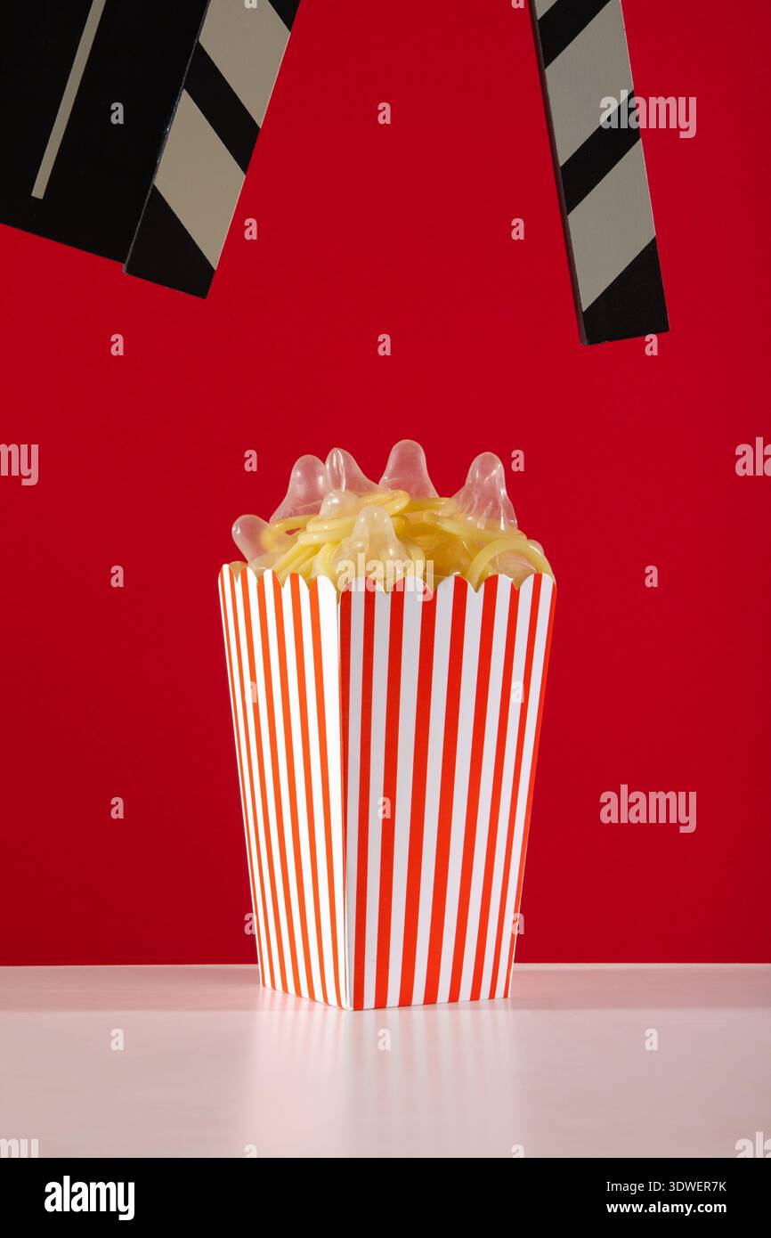 Ein verspieltes und lebendiges Bild mit einer kreativen Popcorn-Box voller farbenfroher verpackter Kondome vor einem kräftigen roten Hintergrund. Ideal für Uniqu Stockfoto