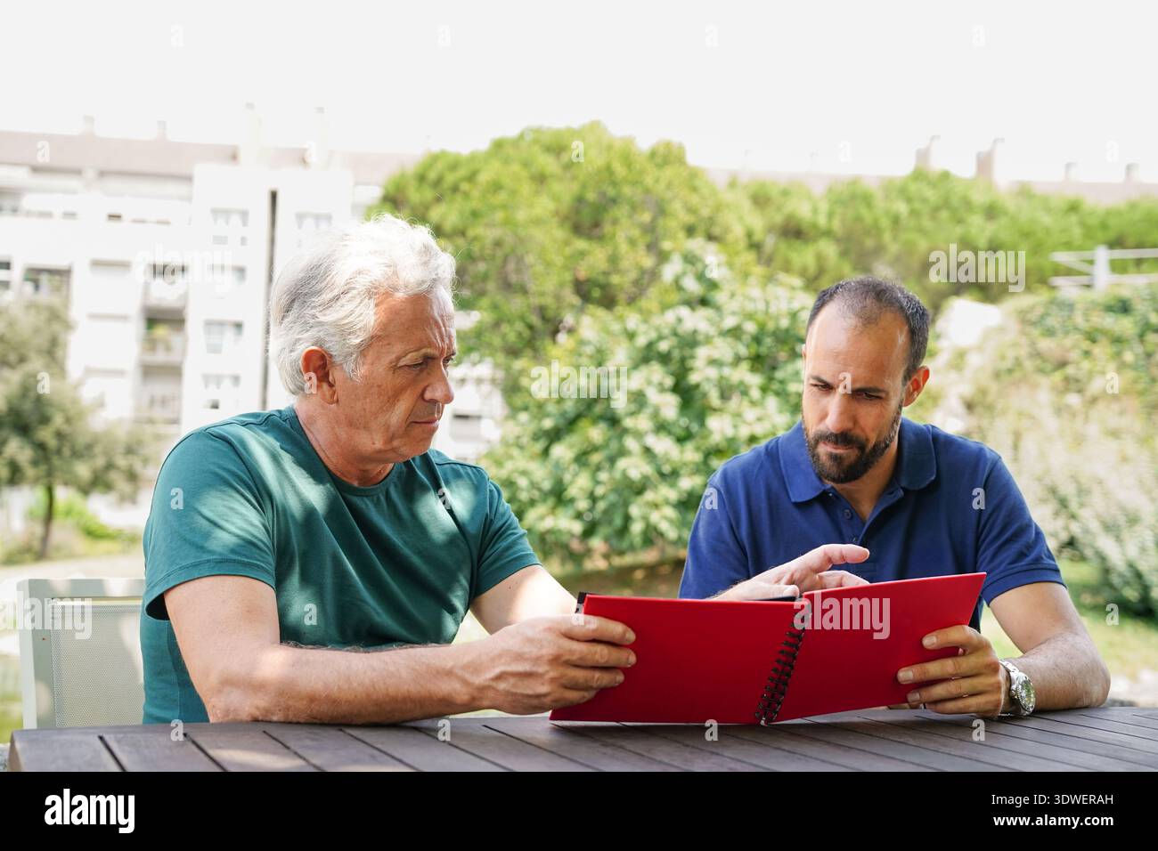Zwei Freunde sitzen draußen an einem Tisch und schauen aufmerksam durch ein rotes Fotoalbum. Der grüne Hintergrund von Bäumen und Gebäuden lässt einen friedlichen Vater vermuten Stockfoto