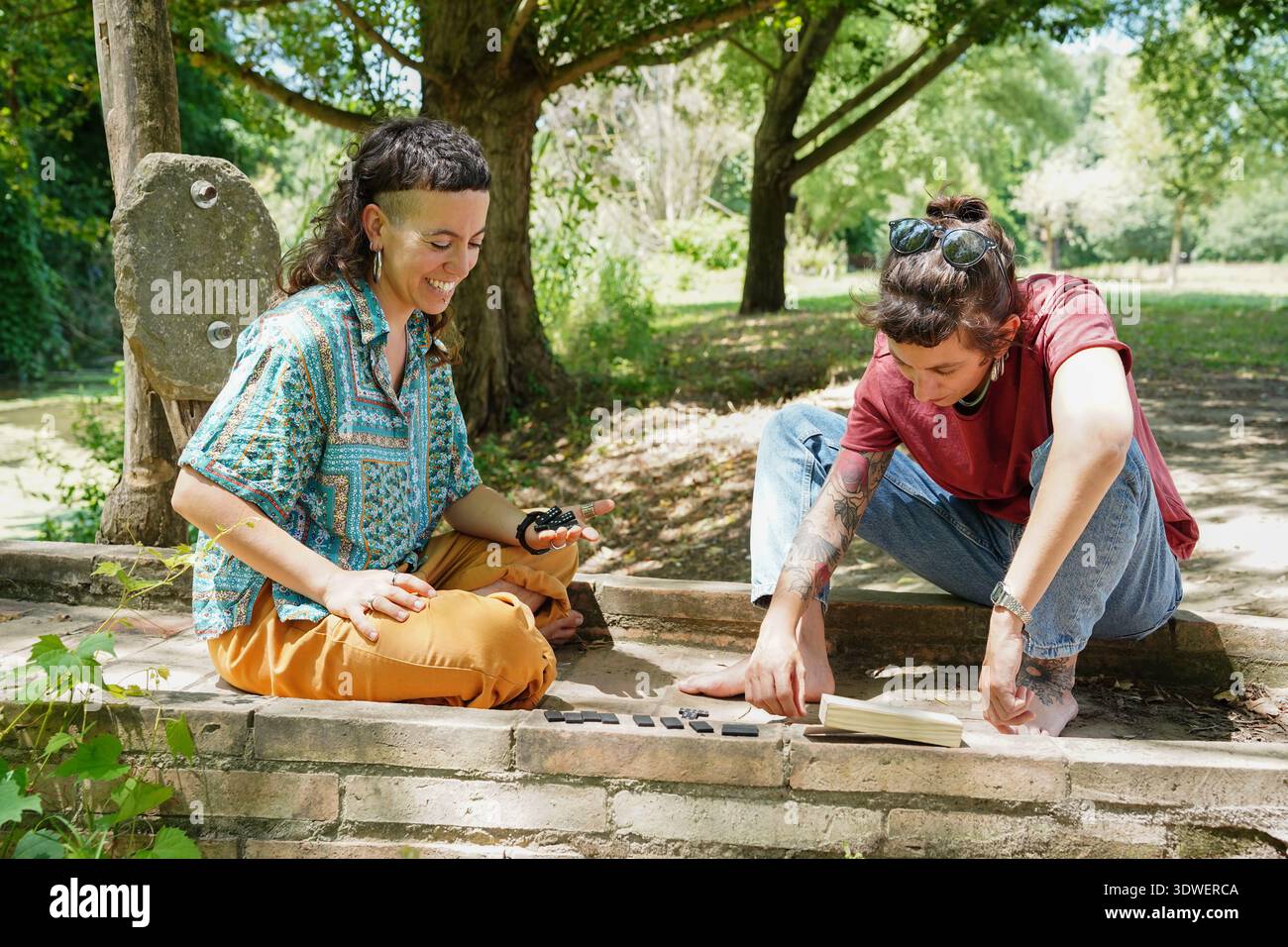 Zwei Schwestern spielen in einem sonnendurchfluteten Park, umgeben von üppigem Grün, ein entspannendes Spiel. Ihr Lächeln und ihre entspannten Posen fesseln das Wesen eines sorglosen Stockfoto
