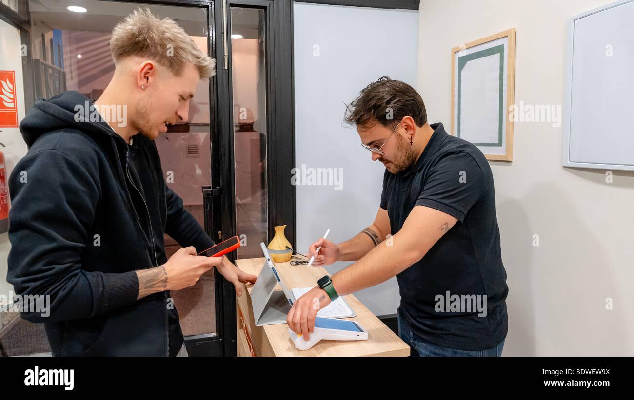 Ein Kunde interagiert mit einem Friseur an der Theke in einem modernen Friseur. Sie scheinen in ein freundliches Gespräch eingebunden zu sein, das eine Begrüßung und p Stockfoto