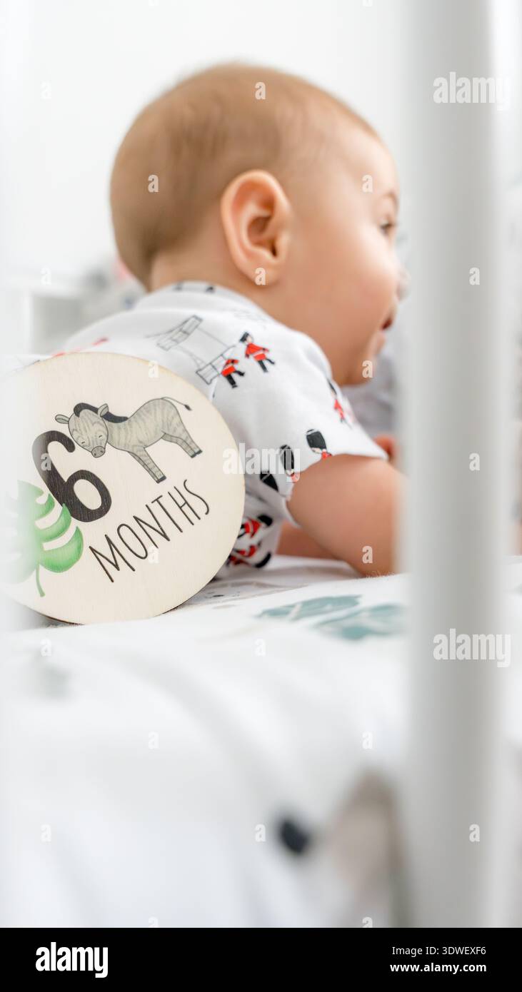 Ein sechs Monate altes Baby liegt auf einem Bett neben einem hölzernen Meilensteinmarker, der 6 Monate anzeigt. Das Baby trägt ein niedliches Outfit, das zum Entspannen beiträgt Stockfoto