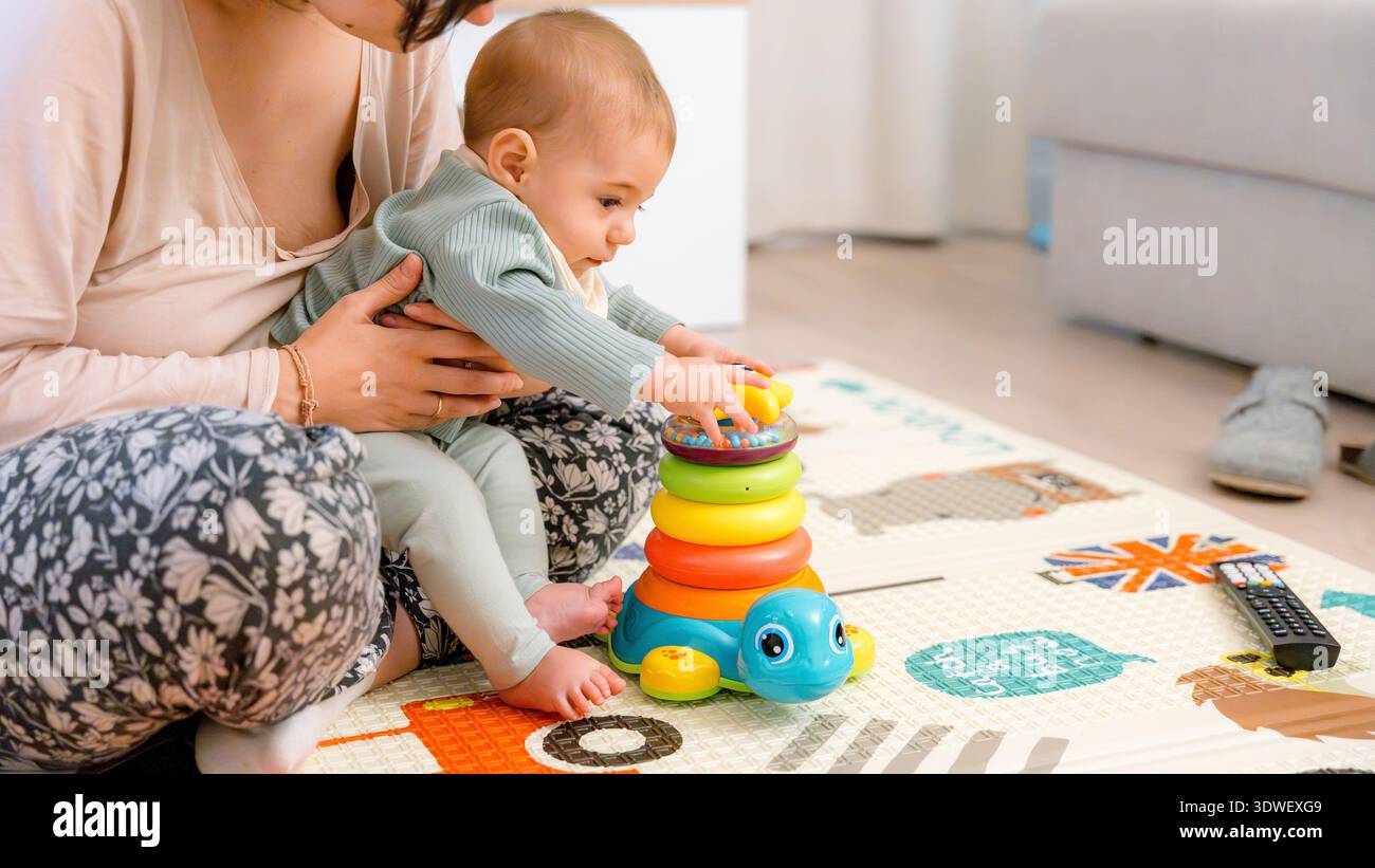 Das Baby sitzt auf einer gemusterten Matte und spielt mit einem bunten, stapelbaren Spielzeug, das von einem Erwachsenen überwacht wird. Die Umgebung ist entspannt, was die frühkindliche P Stockfoto