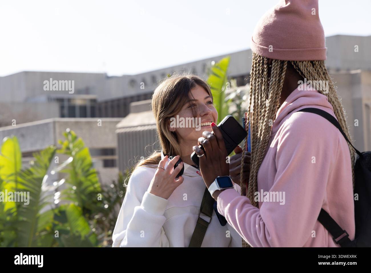 Verschiedene Freundinnen stehen auf dem Campus, halten Smartphone mit Kopfhörern und Rucksack Stockfoto