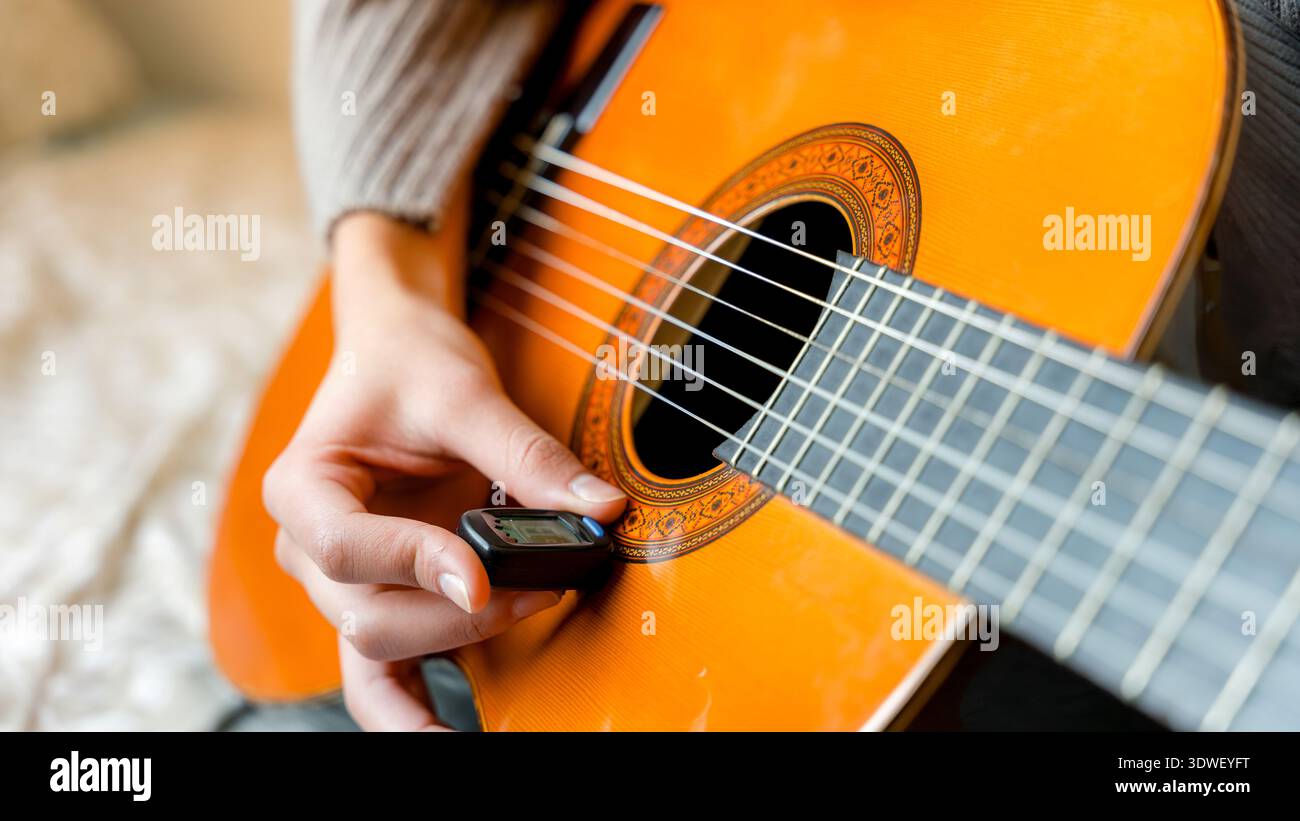 Eine junge Frau stimmt ihre klassische Gitarre sorgfältig mit Präzision ab, wobei sie einen digitalen Tuner verwendet, um eine perfekte Klangqualität zu gewährleisten. Der Fokus liegt auf der Handwerkskunst Stockfoto