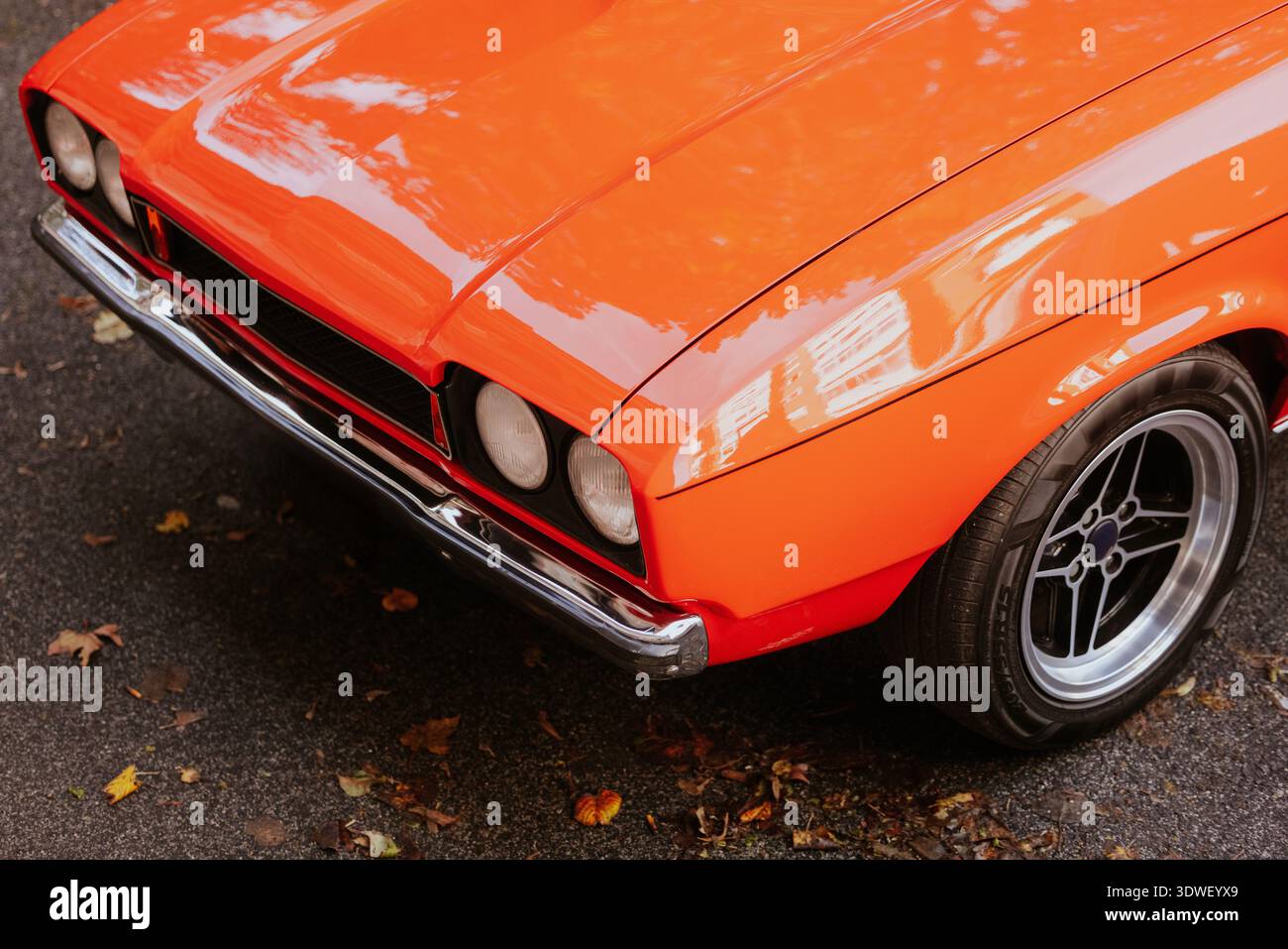 Nahaufnahme eines orangen Oldtimers mit glänzenden Chrom-Details und klassischem Design, der auf einer mit Blättern bedeckten Straße geparkt ist und ein nostalgisches Automotiv erfasst Stockfoto