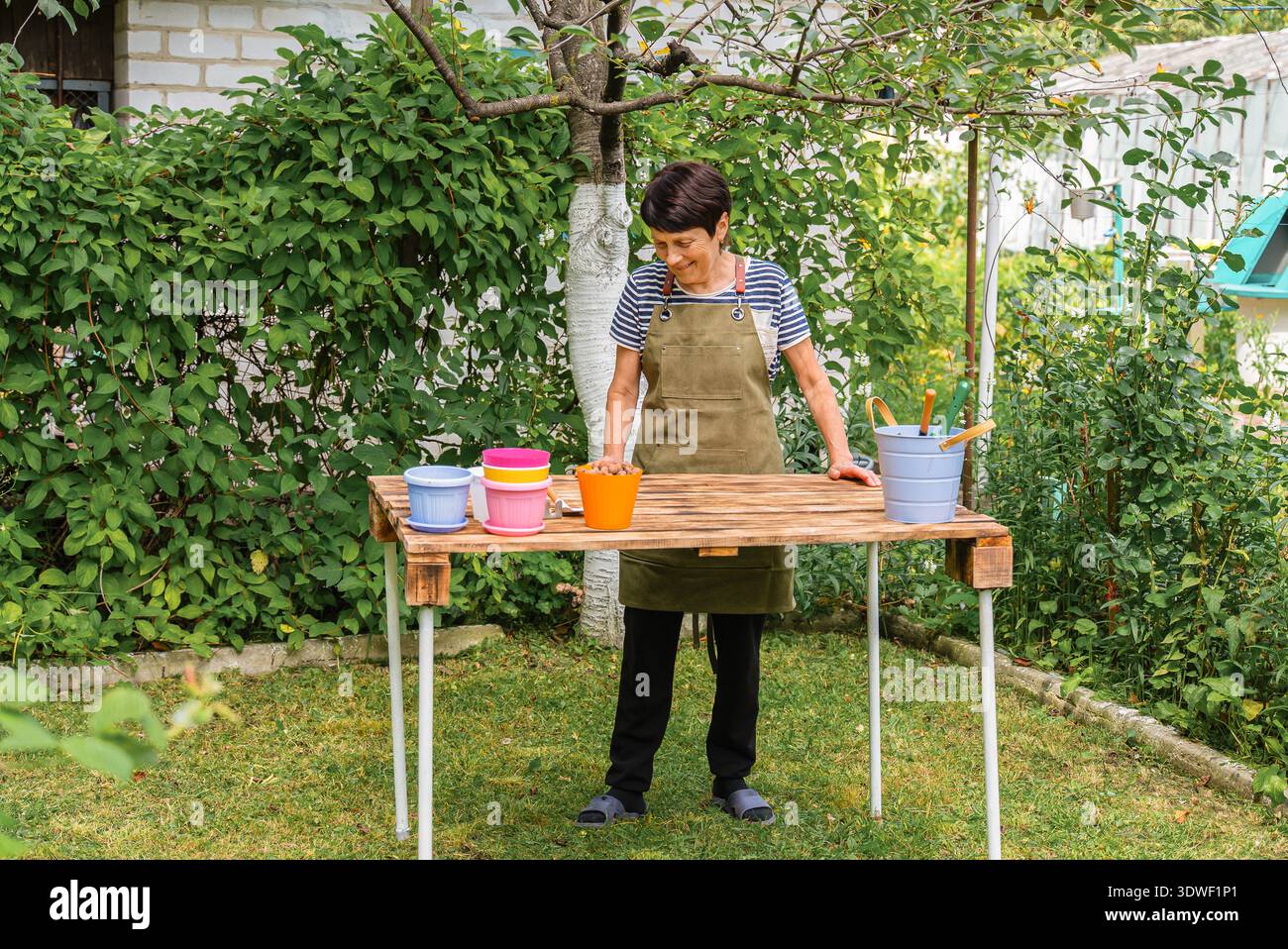 Eine Frau mittleren Alters steht draußen an einem Tisch und bereitet sich darauf vor, Zimmerpflanzen umzupflanzen. Sie trägt eine Schürze und ist von Gartenwerkzeugen umgeben und farbenfroh Stockfoto