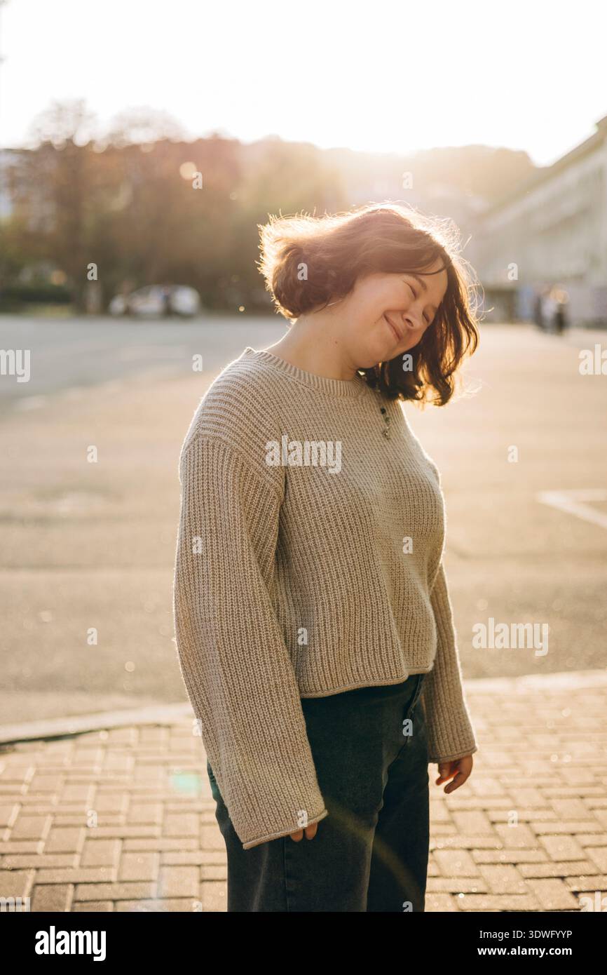 Eine Frau steht draußen in einem lässigen beigefarbenen Pullover und genießt die warme Sonne. Das entspannte und ruhige Ambiente wird durch das goldene Hell ca. Stockfoto