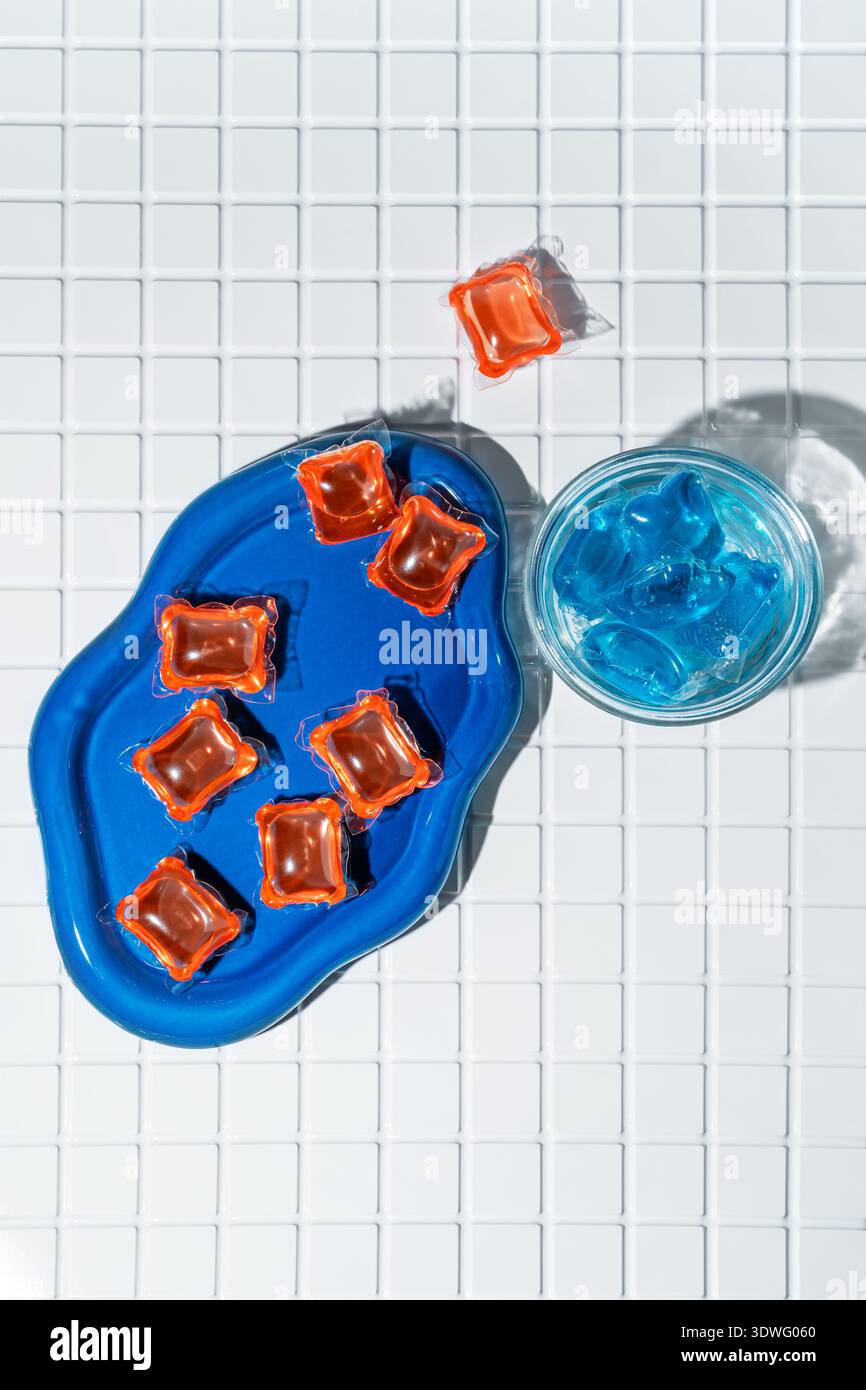 Leuchtend orange und blaue Waschkapseln ruhen auf einer blauen Schale vor einem weißen gefliesten Hintergrund. Der Kontrast unterstreicht die lebendigen Farben und der Kapseln Stockfoto