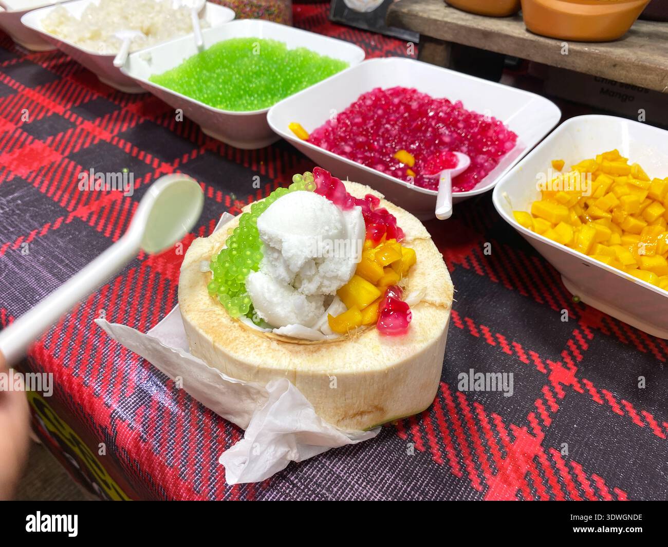 Kokos-Eis in einer Kokosnussschale mit lebhaften Belägen wie Obst und Gelee, auf eine karierte Tischdecke mit Schüsseln mit zusätzlichen Belägen ne gelegt Stockfoto