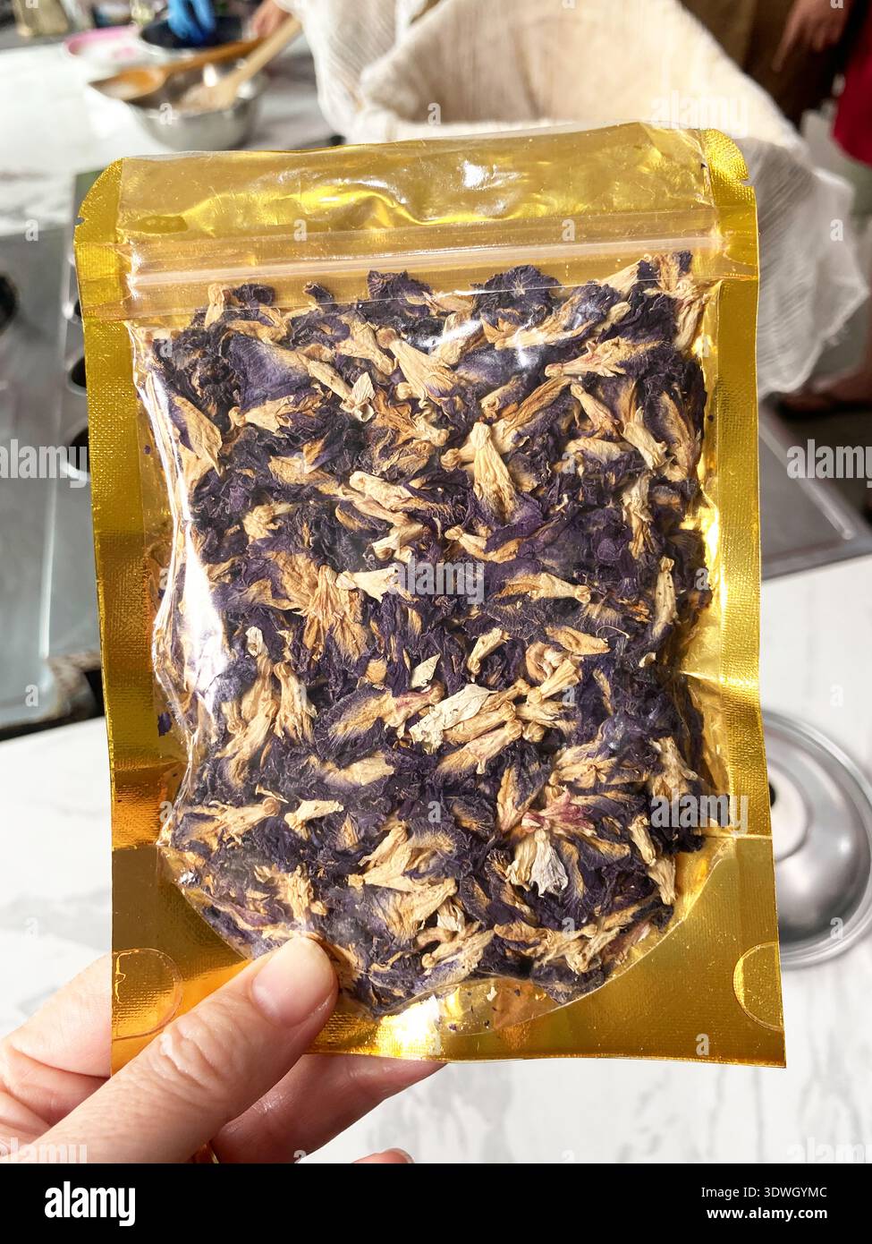 Getrocknete Schmetterlingserbsenblüten werden in einem transparenten Goldpaket dargestellt, das von einer Hand gehalten wird, wobei eine Kücheneinrichtung im Hintergrund sichtbar ist Stockfoto