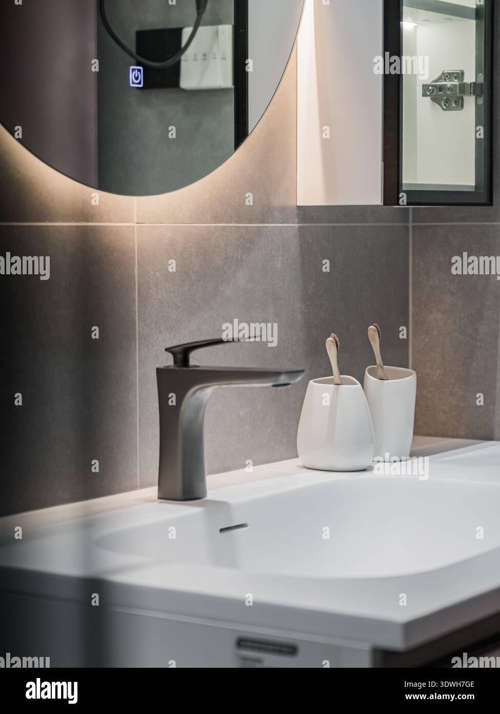 Das Badezimmer verfügt über ein elegantes Waschbecken mit modernem Wasserhahn. Neben dem Wasserhahn befinden sich zwei kleine dekorative Behälter und ein runder Spiegel über dem Si Stockfoto