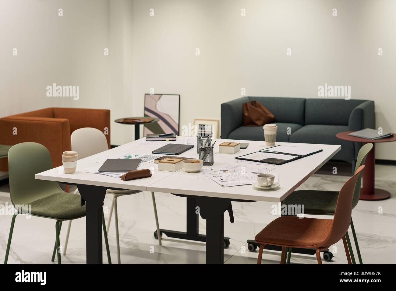 Moderner Büroarbeitsplatz mit leerem Tisch mit verstreuten Dokumenten, Kaffeetassen, digitalen Tablets, Schreibwaren und Stühlen, Hintergrund mit Sofabühlen und minimalistischem Dekor Stockfoto
