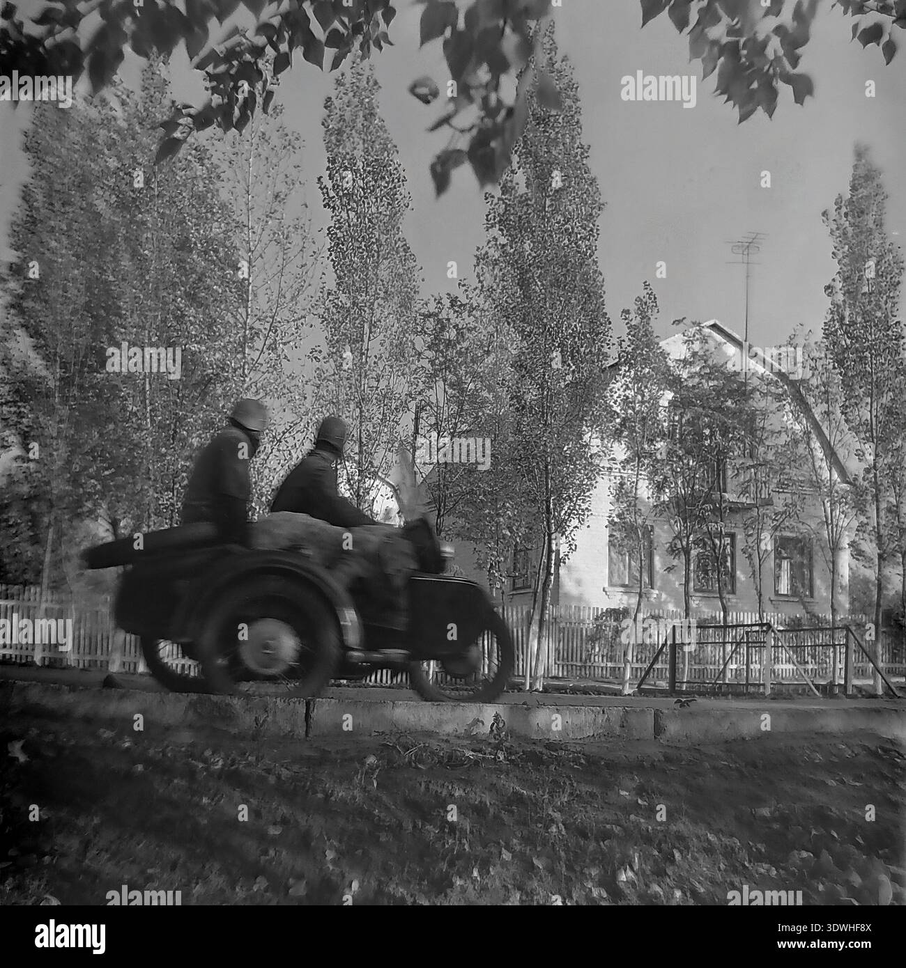 Das historische Schwarzweißfoto aus den 1970er Jahren zeigt ein klassisches sowjetisches schweres Motorrad mit einem Beiwagen, der auf einer ruhigen Feldstraße in der Nähe des Donrybkombinats im Bezirk Slavjansk, ukrainische SSR, UdSSR, fährt. Zwei Helmfahrer fahren an einem Holzzaun, hohen Bäumen und einem zweistöckigen Backsteinhaus vorbei. Dieses authentische Archivbild zeigt die Dynamik des ländlichen Verkehrs, der alltäglichen Dorfinfrastruktur und des stabilen Alltags im friedlichen Donbass vor dem Krieg und spiegelt die regionale Mobilität wider. Stockfoto