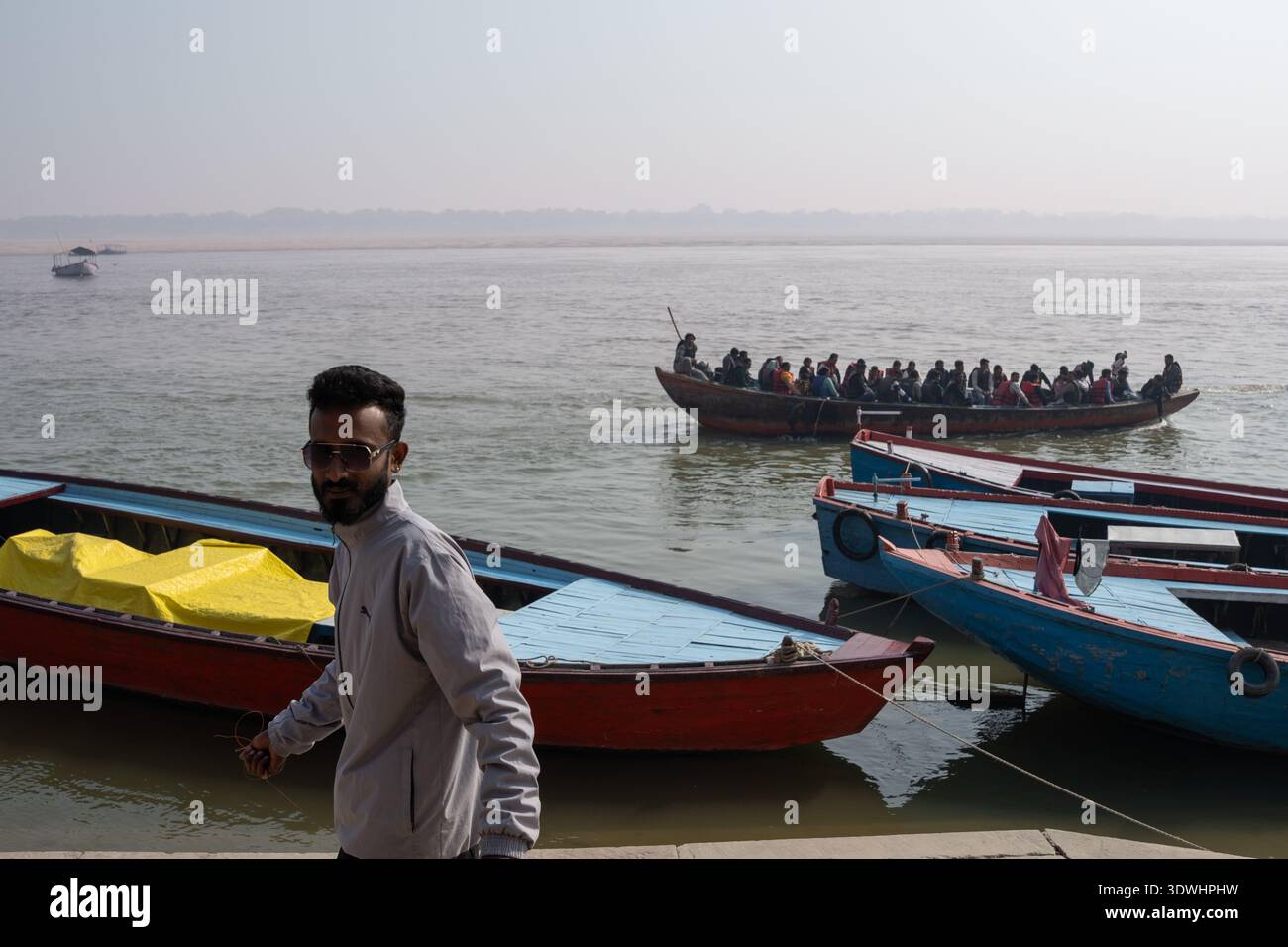 Überfüllte Boote auf dem Fluss in Varanasi, Indien, mit einem Mann, der Sonnenbrille trug, im Vordergrund in der Nähe des Ufers. Stockfoto