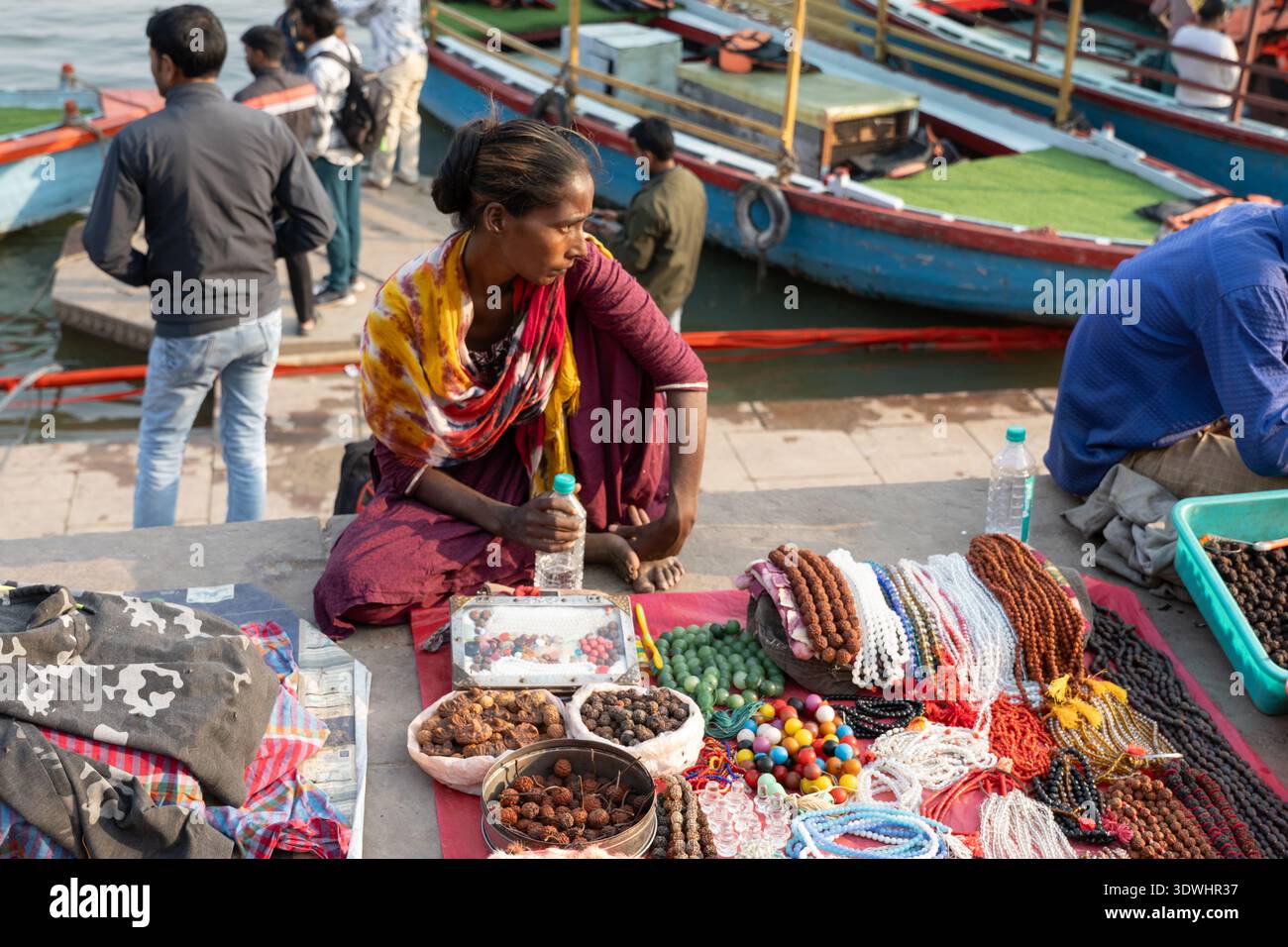 Eine Frau, die bunte Perlen und Schmuck auf einem Markt am Flussufer in Varanasi, Indien, verkauft, mit Booten im Hintergrund. Stockfoto