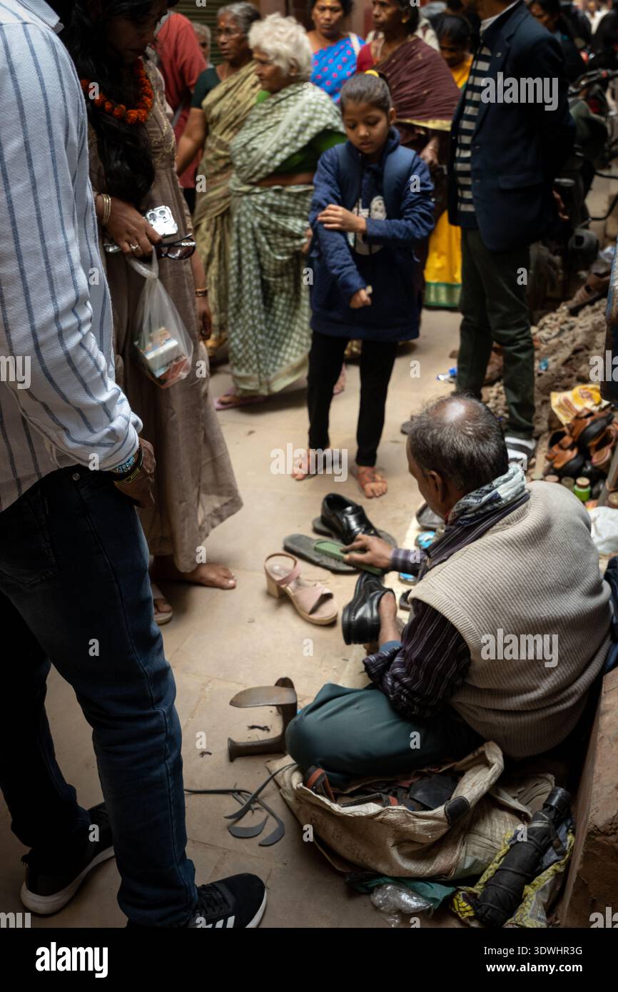 Ein Schuster repariert Schuhe auf einer geschäftigen Straße in Varanasi, Indien, umgeben von Einheimischen und traditioneller Kleidung. Stockfoto