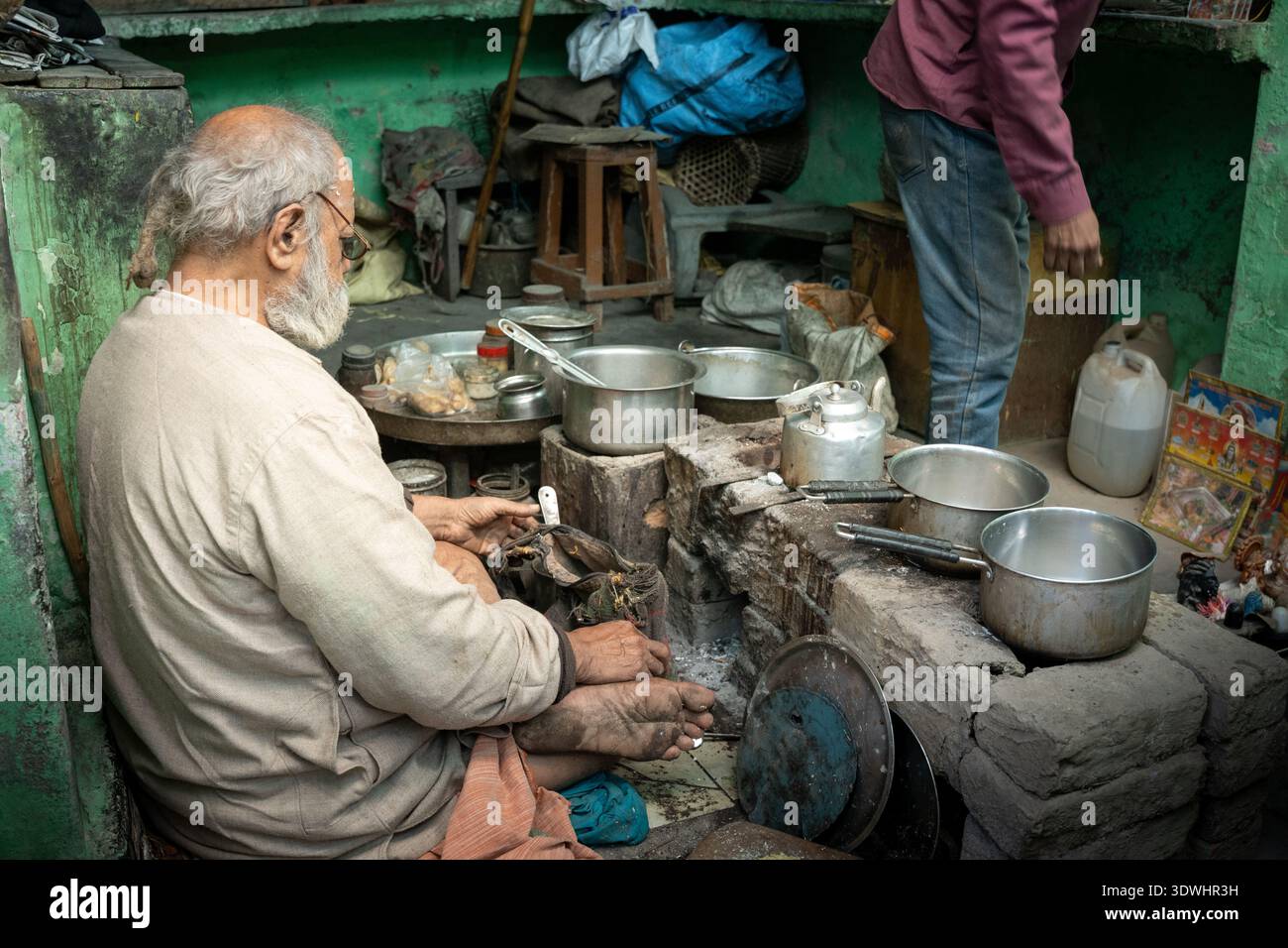 Ein älterer Mann, der Milchtee in einer rustikalen Küche in Varanasi, Indien, zubereitet, umgeben von Kochtöpfen und Utensilien. Stockfoto