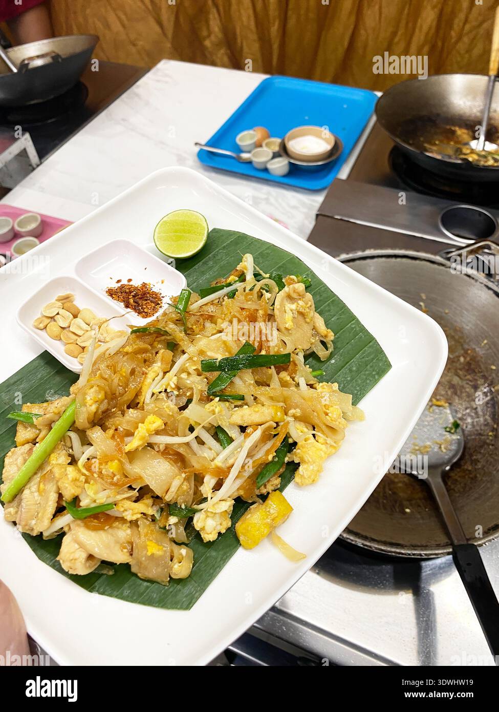 Teller mit Pad thai mit Huhn, Sojasprossen und Limette auf einem Bananenblatt, mit Gewürzen und Kochutensilien im Hintergrund Stockfoto