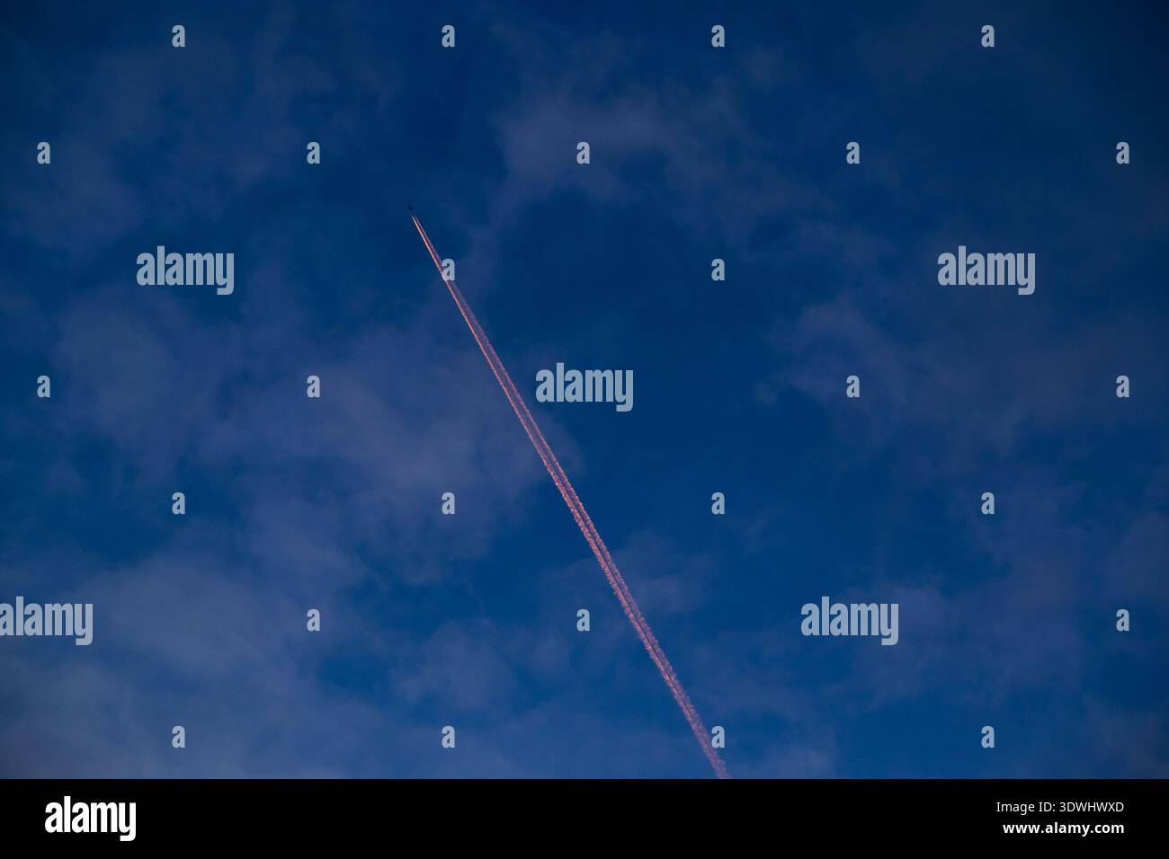 Leuchtend orangefarbener Kondensstreifen teilt einen tiefblauen Abendhimmel mit weichen Wolken Stockfoto