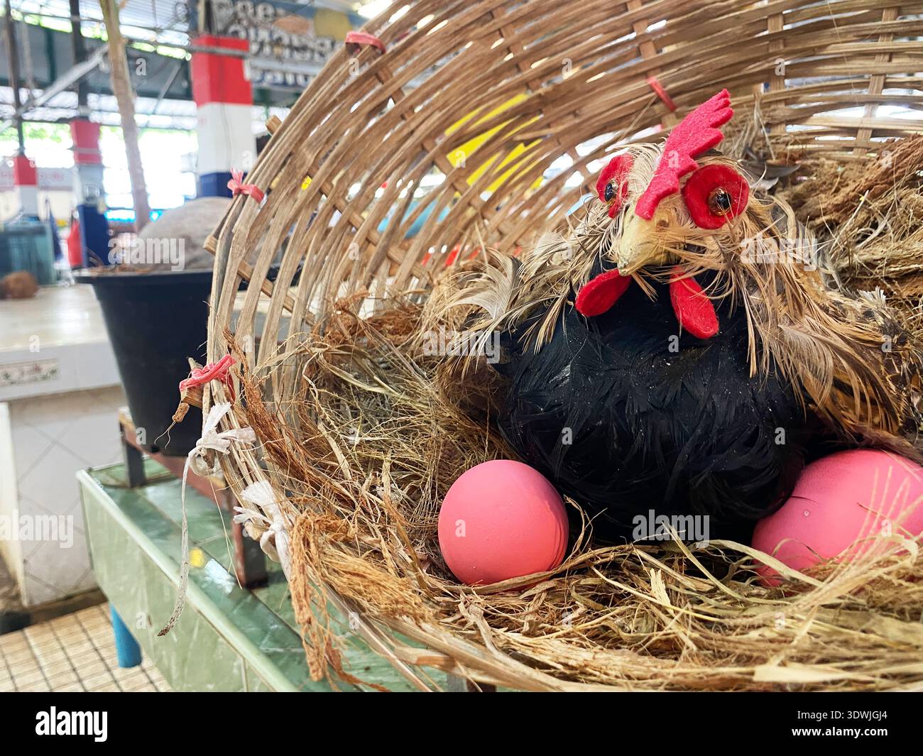 Hühnerpuppe mit dekorativen Elementen und rosa Eiern in einem gewebten Korb auf einem Markttisch, umgeben von einer lebhaften Marktkulisse Stockfoto