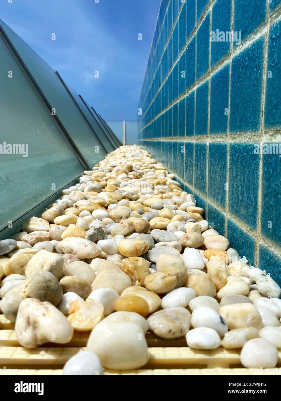 Dekorativer Kiesweg mit glatten Kieselsteinen entlang einer blau gekachelten Wand, mit klarem Himmel und modernen architektonischen Elementen in einem Pool Stockfoto