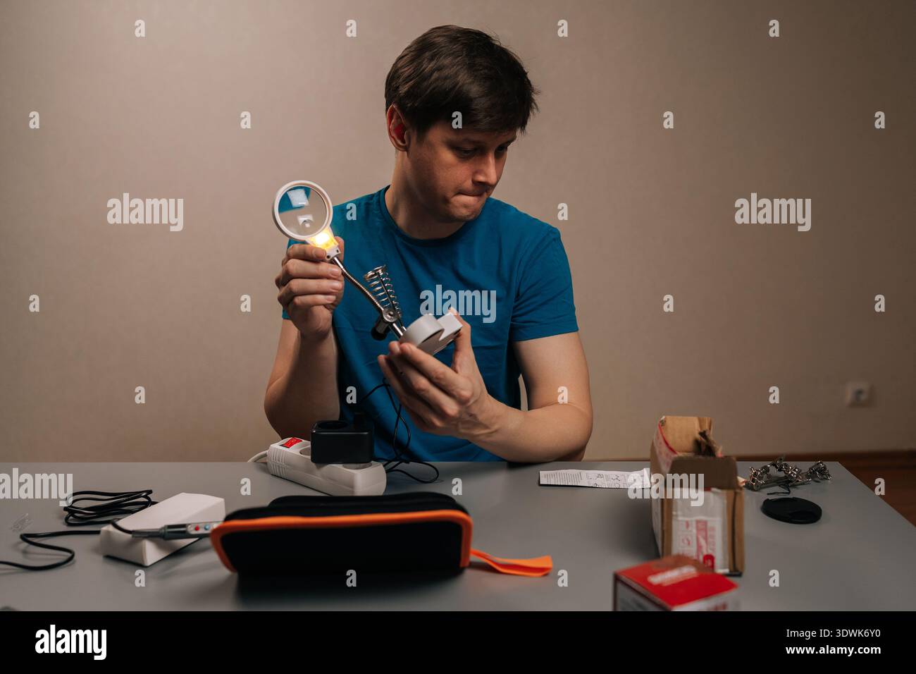 Mann, der sich auf ein elektronisches Gerät konzentriert, Komponenten mit einer beleuchteten Lupe überprüft, während er sich auf das Löten vorbereitet, und Präzision in einem Zuhause demonstriert Stockfoto