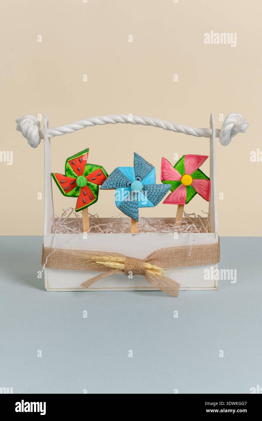Drei leuchtende Räderchen in einem weißen Korb mit Seilgriff, farbenfrohe Designs, die auf einer hellblauen Oberfläche vor einem beigefarbenen Hintergrund platziert sind Stockfoto
