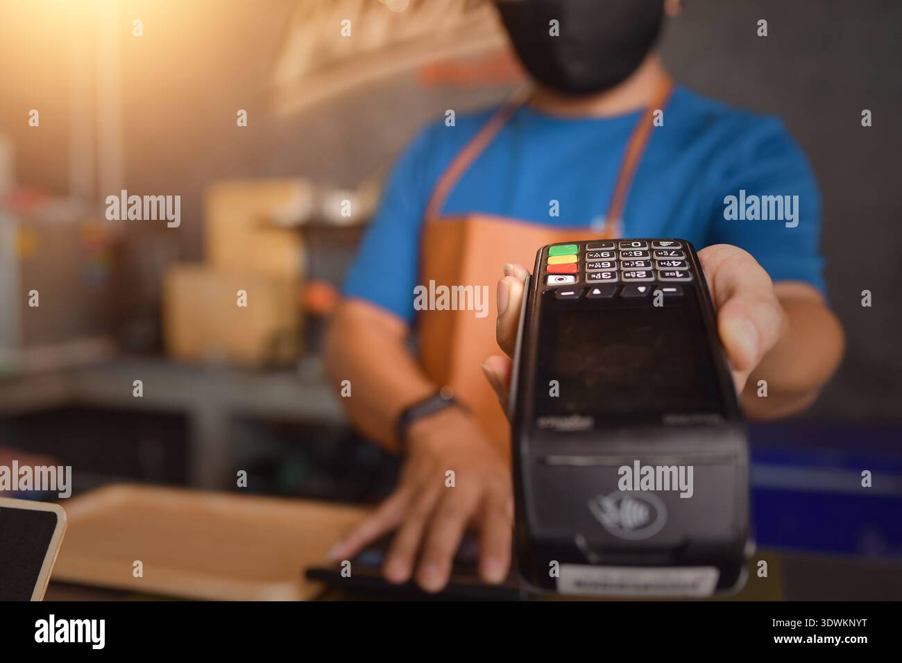 Nahaufnahme eines Verkäufers mit Schürze und Gesichtsmaske, der in einem Geschäft steht, das ein Kreditkartenzahlungsterminal hält Stockfoto
