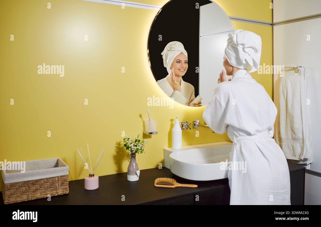 Glückliche, schöne Frau, die in den modernen Badezimmerspiegel blickte und einen weißen Bademantel und ein Handtuch trug Stockfoto