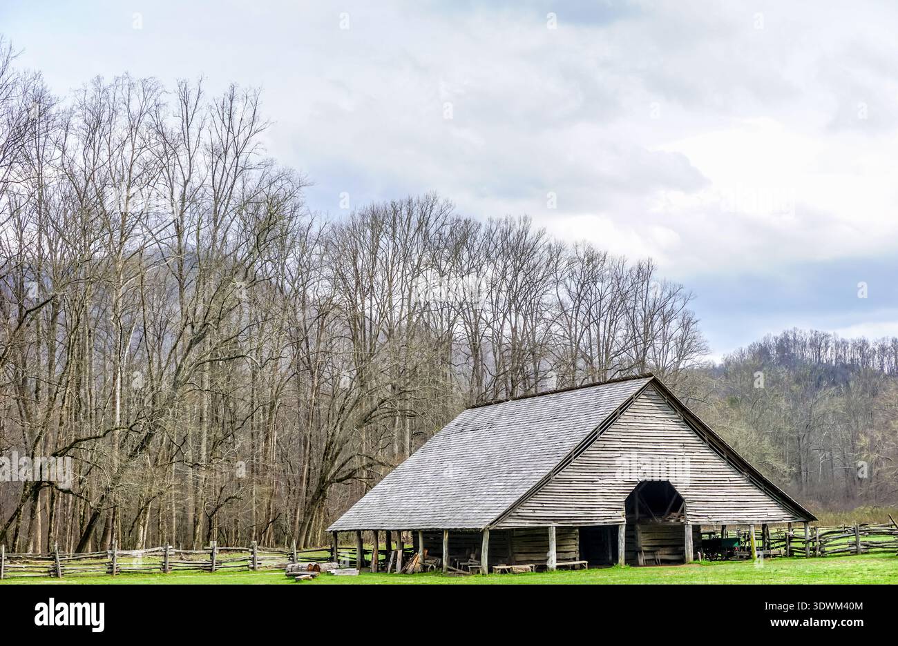Eine große, alte Scheune sitzt auf einem Feld mit Bäumen im Hintergrund. Die Scheune ist von einem Zaun umgeben und er befindet sich in einer ländlichen Umgebung Stockfoto