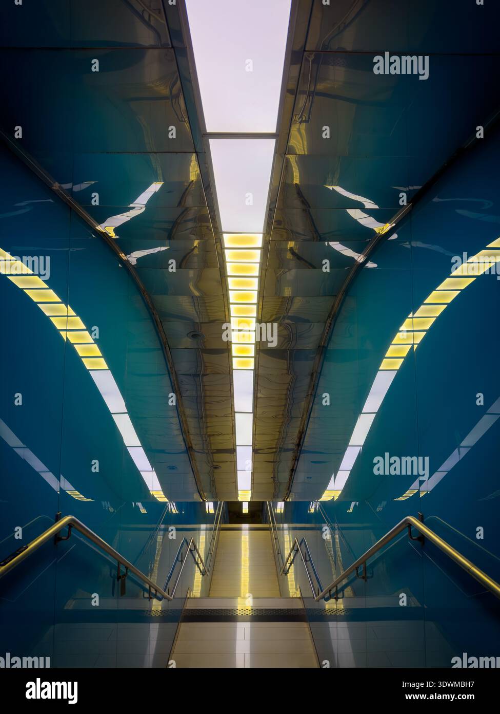 Moderne unterirdische Stationstreppe mit glänzend blauen Wänden, symmetrischen Geländern und warmen linearen Lichtern, die futuristische Stimmung und starke Perspektive schaffen. Stockfoto