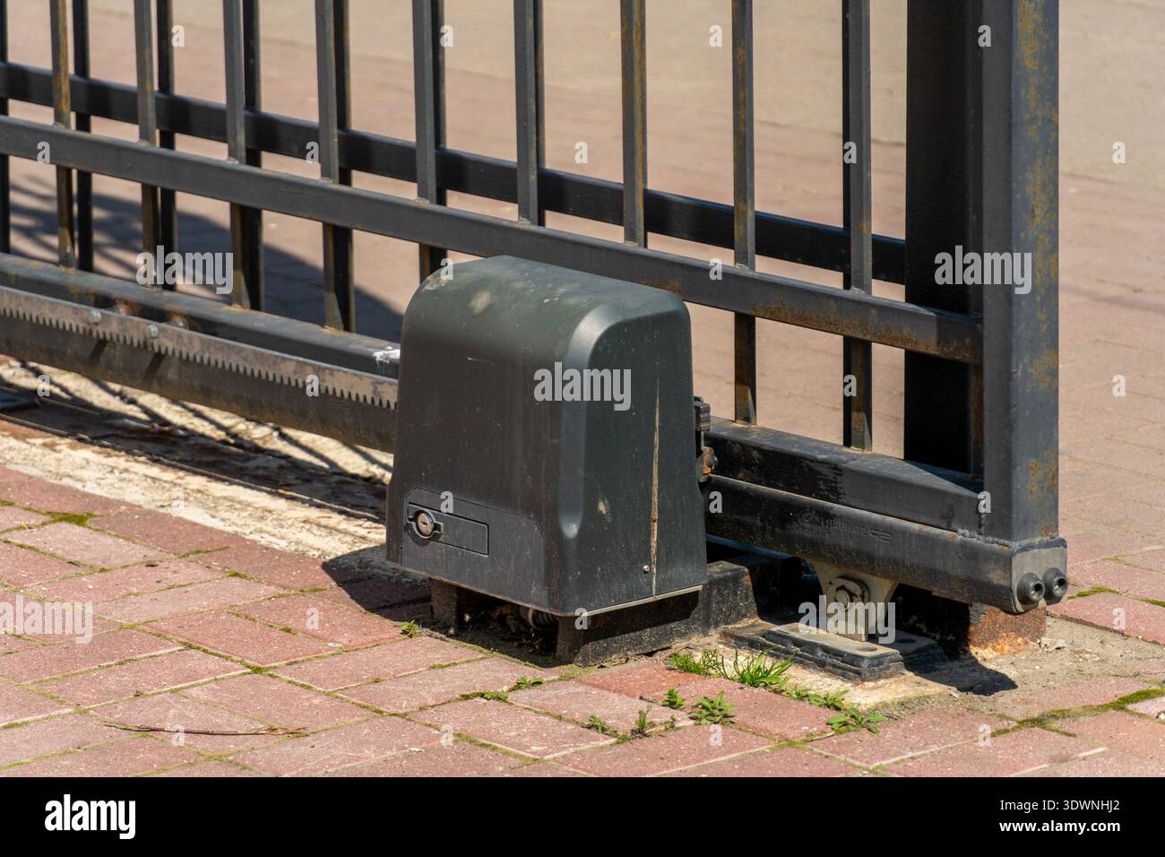 Ein Elektroschiebermotor befindet sich auf dem Boden neben einem Metallzaun. Das Tor ist geöffnet. Die Einstellung ist bei Tageslicht im Freien. Stockfoto