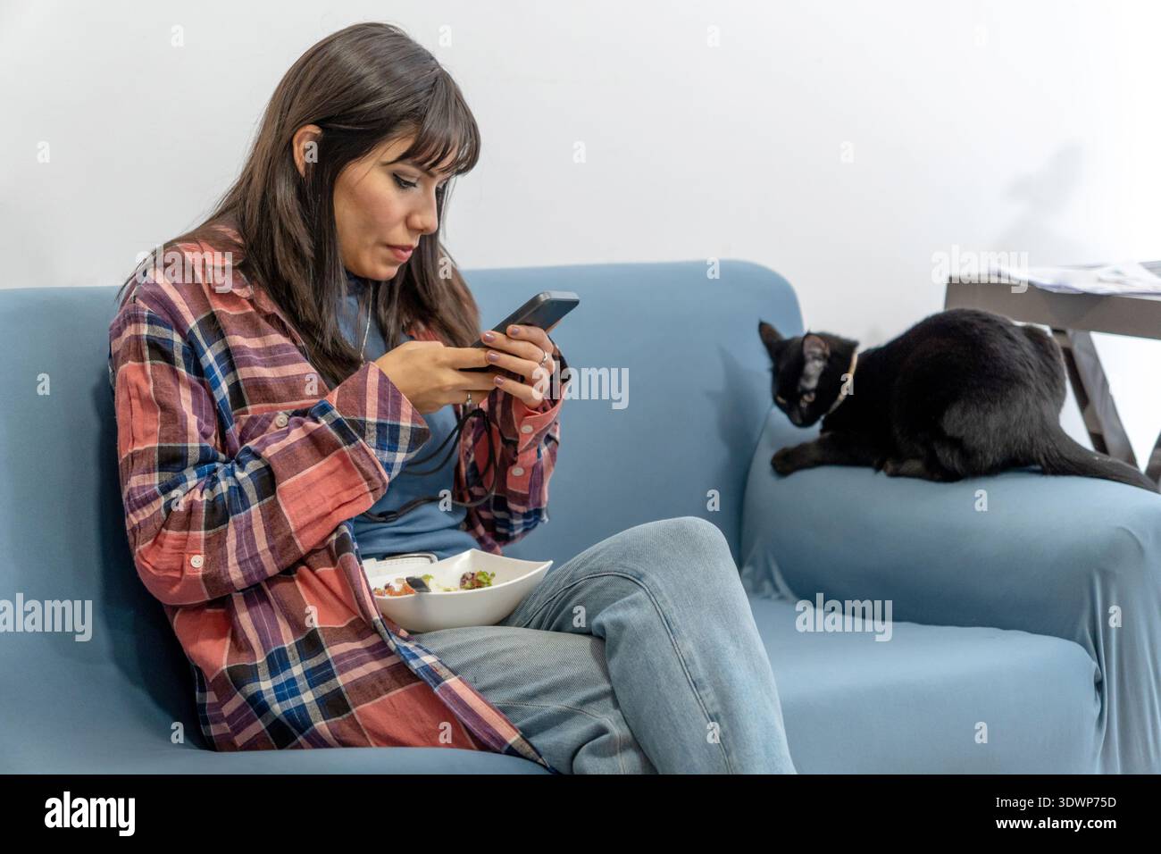 Frau, die sich auf der Couch entspannt und ein gesundes Salat mit einer schwarzen Katze neben ihr isst Stockfoto