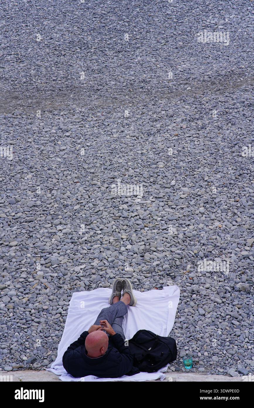 Glatze Mann, der sich auf einem weißen Handtuch am Kiesstrand in Nizza, Frankreich, mit Händen hinter dem Kopf unter einem milden Winterhimmel entspannt. Nizza, Frankreich Stockfoto