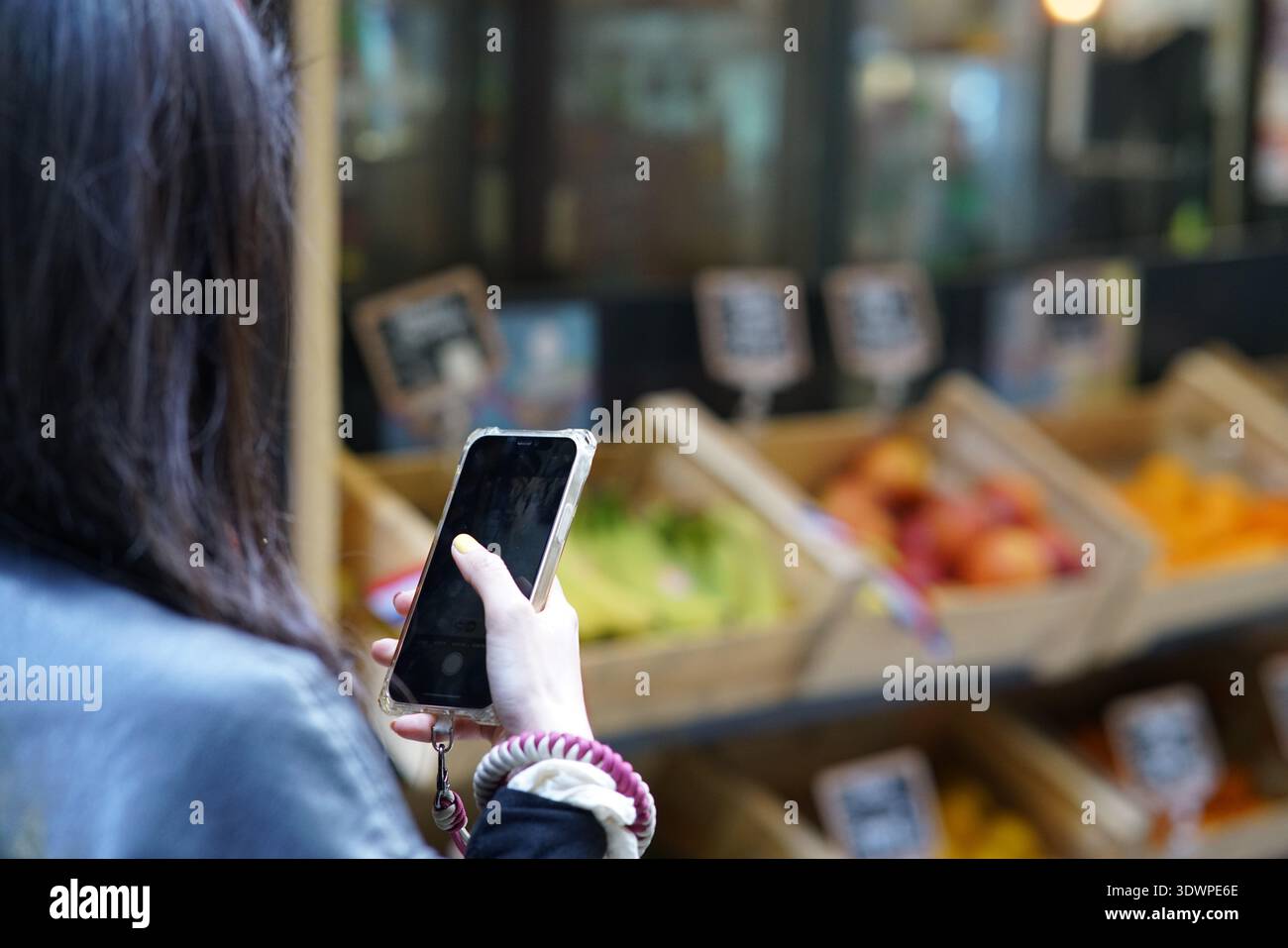 Eine junge Frau in blauer Jacke fängt auf einem Markt in Nizza auf ihrem Smartphone einen lebhaften Obststand voller Bananen, Äpfel, Orangen und Pfirsiche ein. Stockfoto