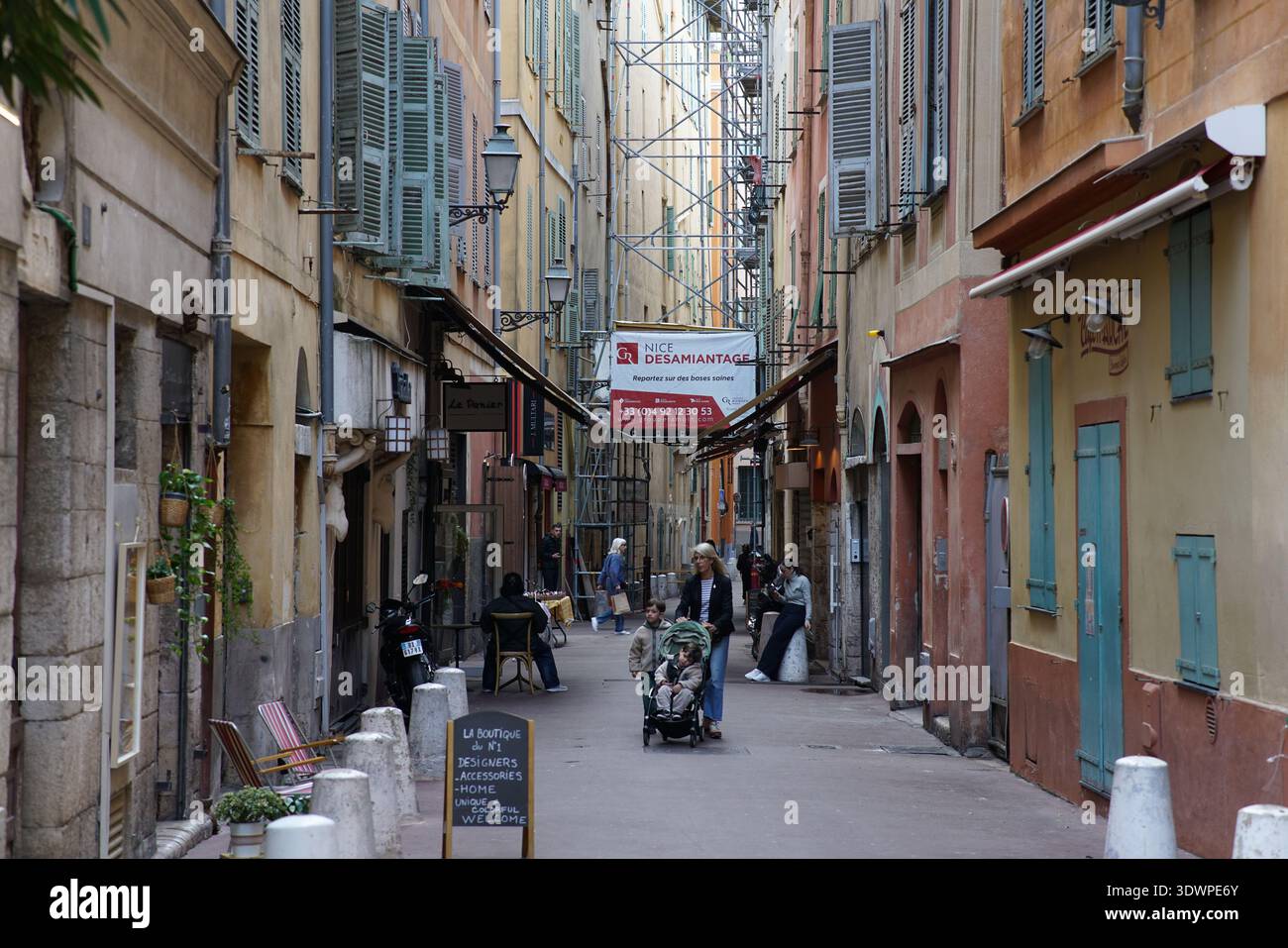 Enge, farbenfrohe Gasse in Vieux Nice mit überhängender Fahne, pastellfarbenen Fassaden, grünen Fensterläden, Spaziergänger und Cafés im Freien Stockfoto